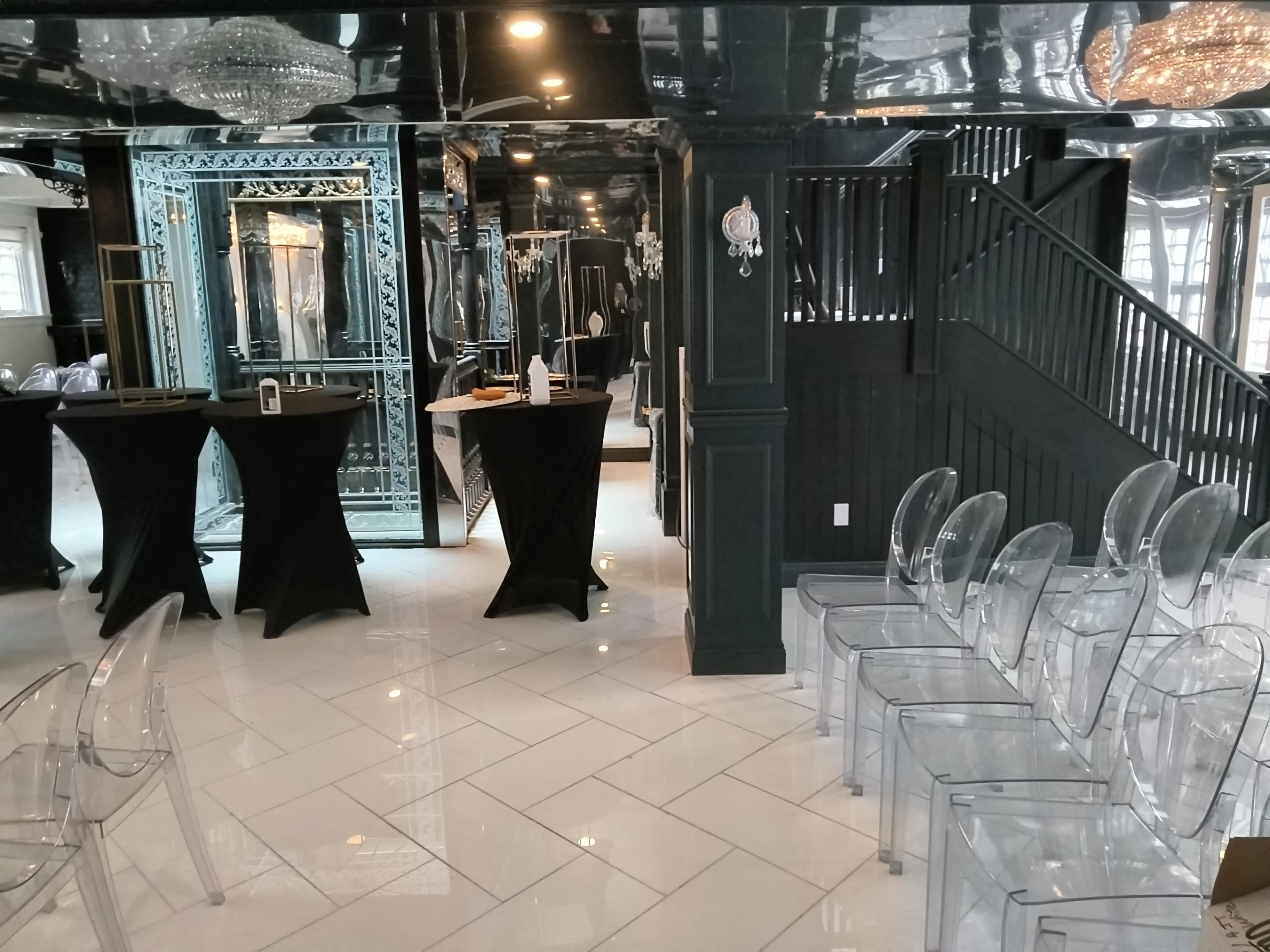 The image shows a stylishly decorated indoor space featuring black and white decor, with tall black tables and transparent chairs arranged for an event, along with a staircase and mirrors on the walls.