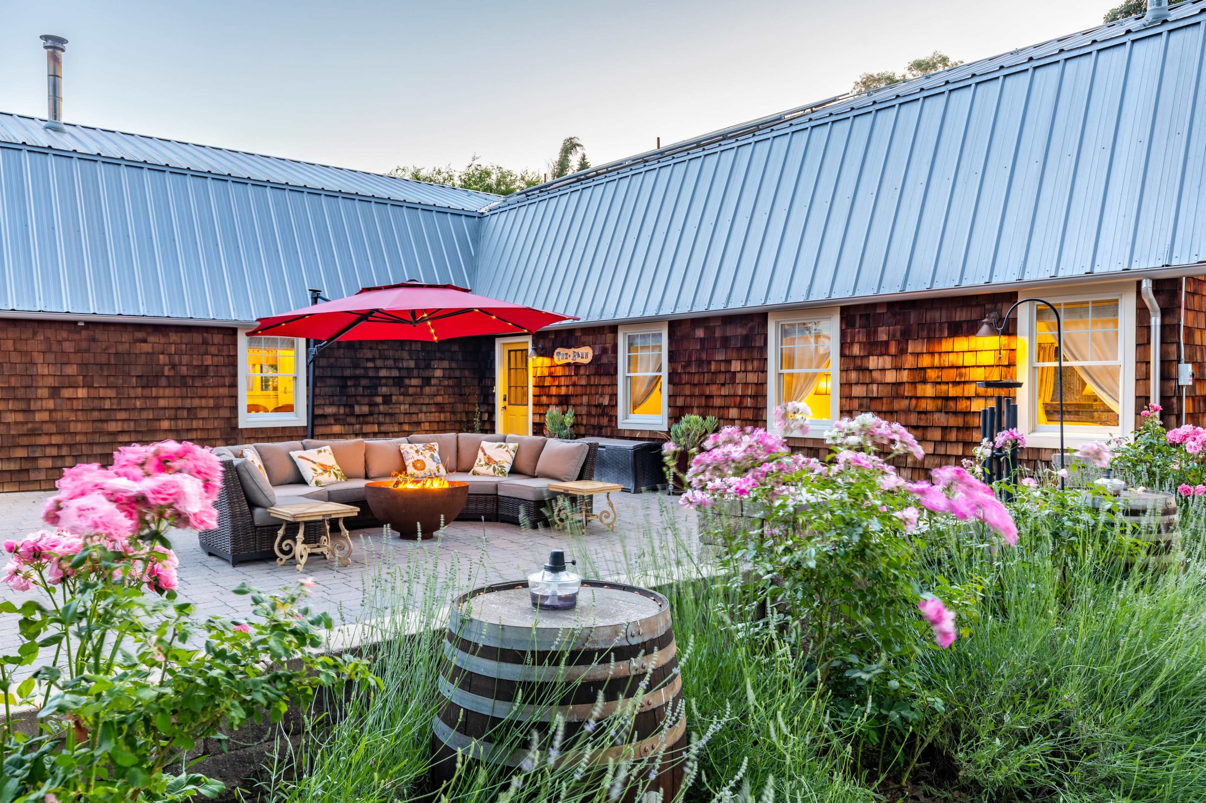 A cozy outdoor seating area with a fire pit is surrounded by flowers and rustic wooden barrels in a courtyard.