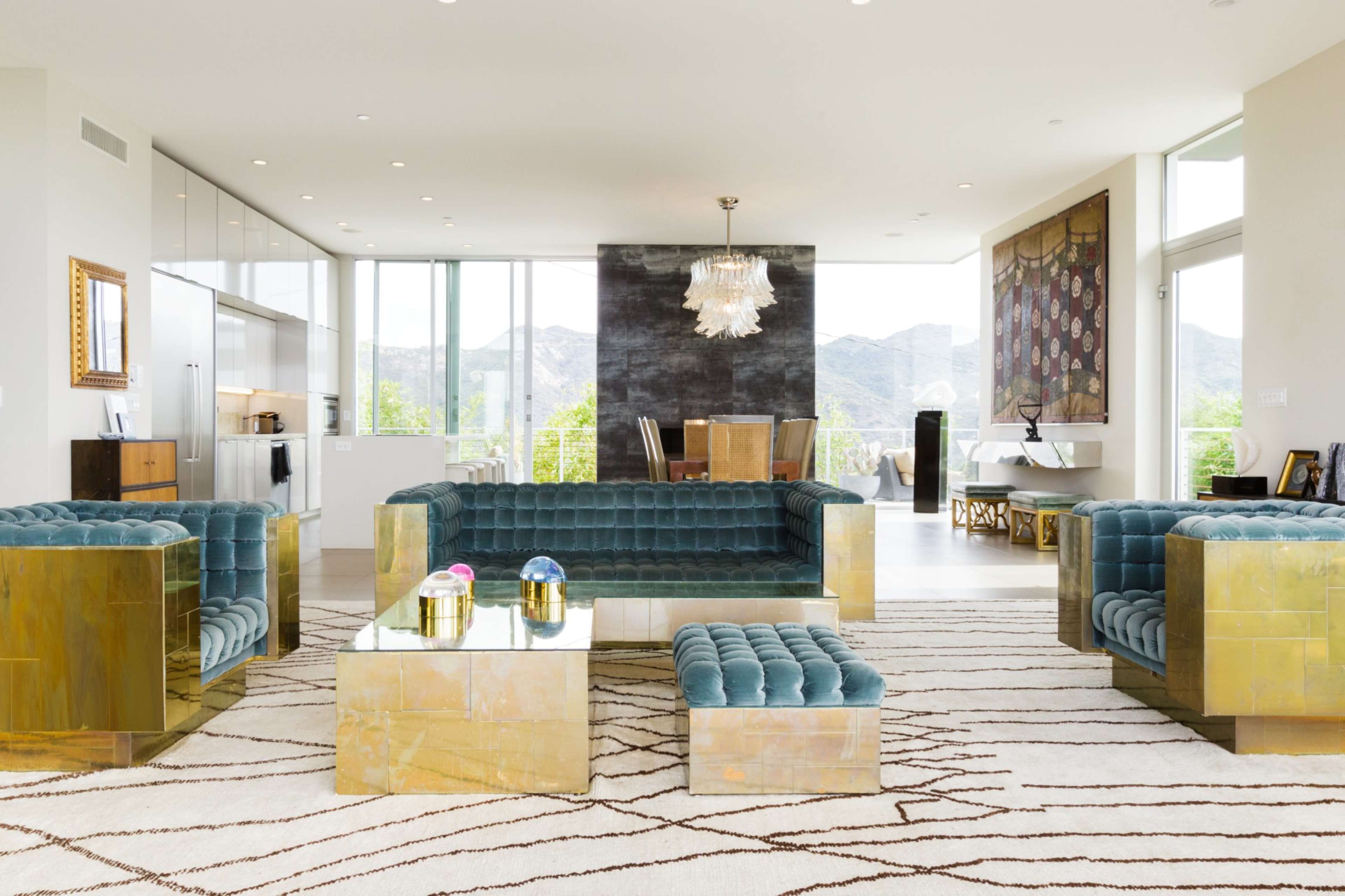 A contemporary living room with teal upholstered couches and gold-accented furniture, overlooking a scenic view through large windows.