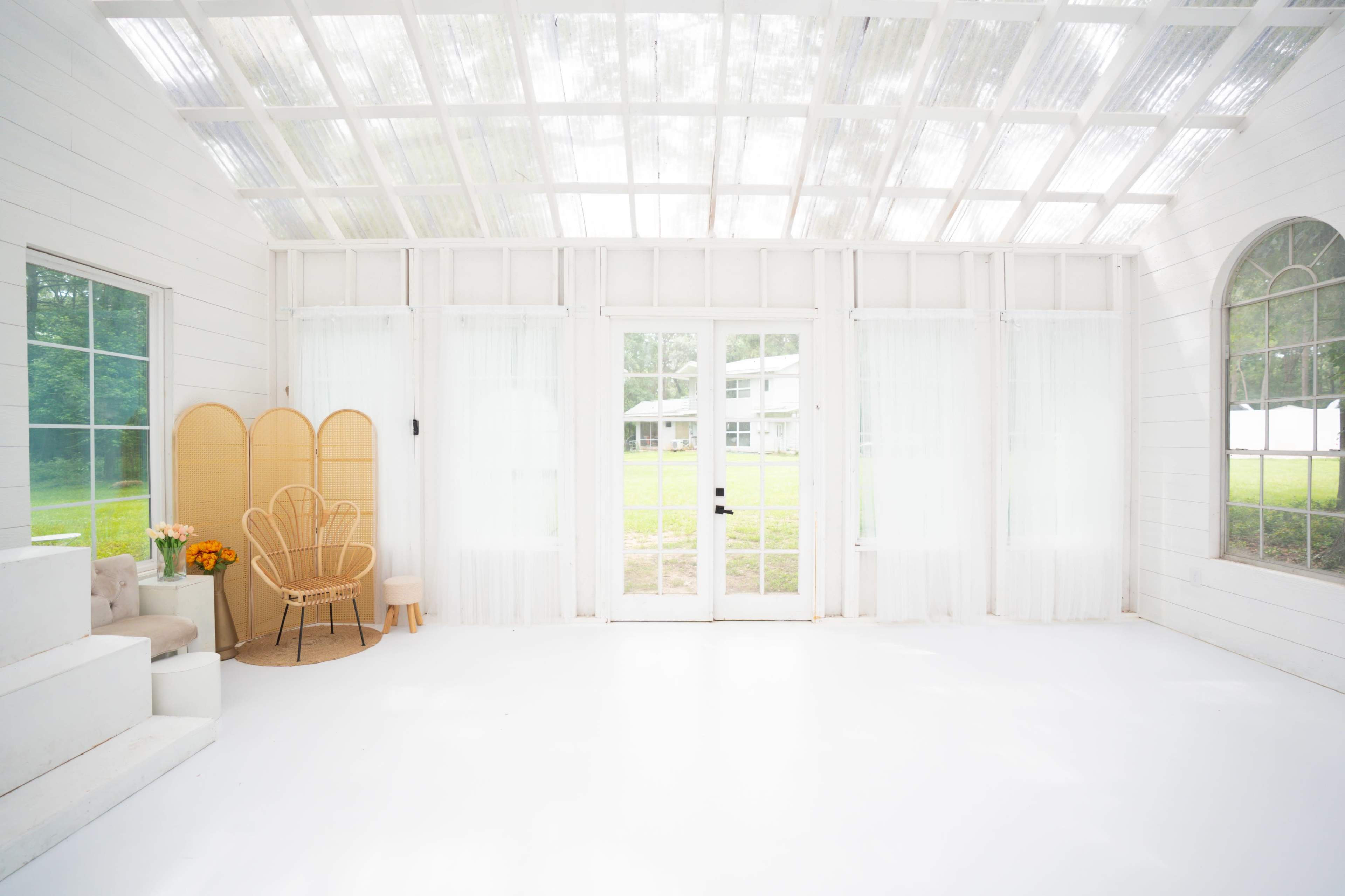 The interior of a bright, airy room features large windows, white walls, and a minimalist design with a seating area and potted flowers.