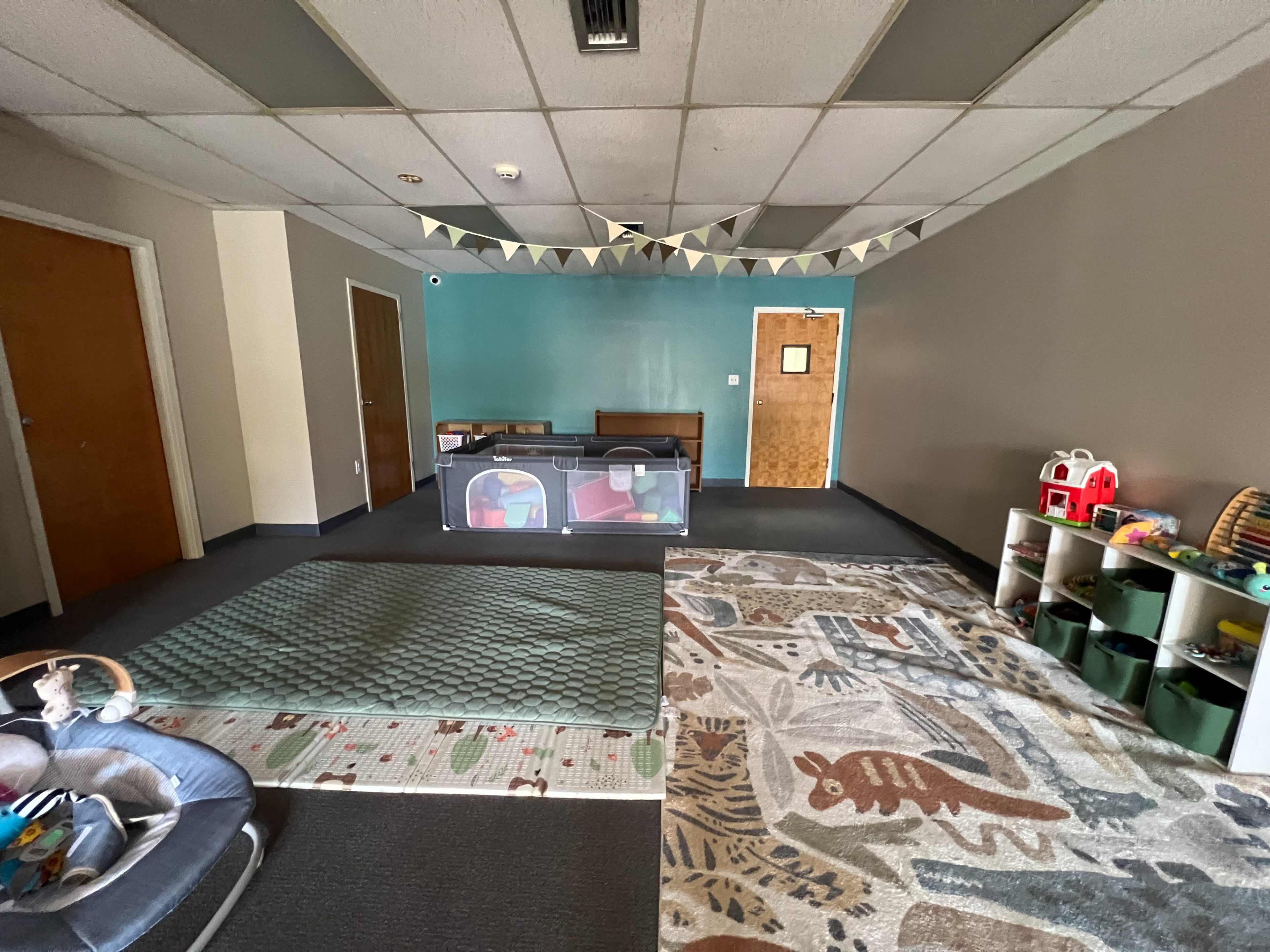 The image shows an empty playroom featuring a large area rug, a soft play mat, a partitioned playpen, storage bins, and a few toys scattered across the floor.