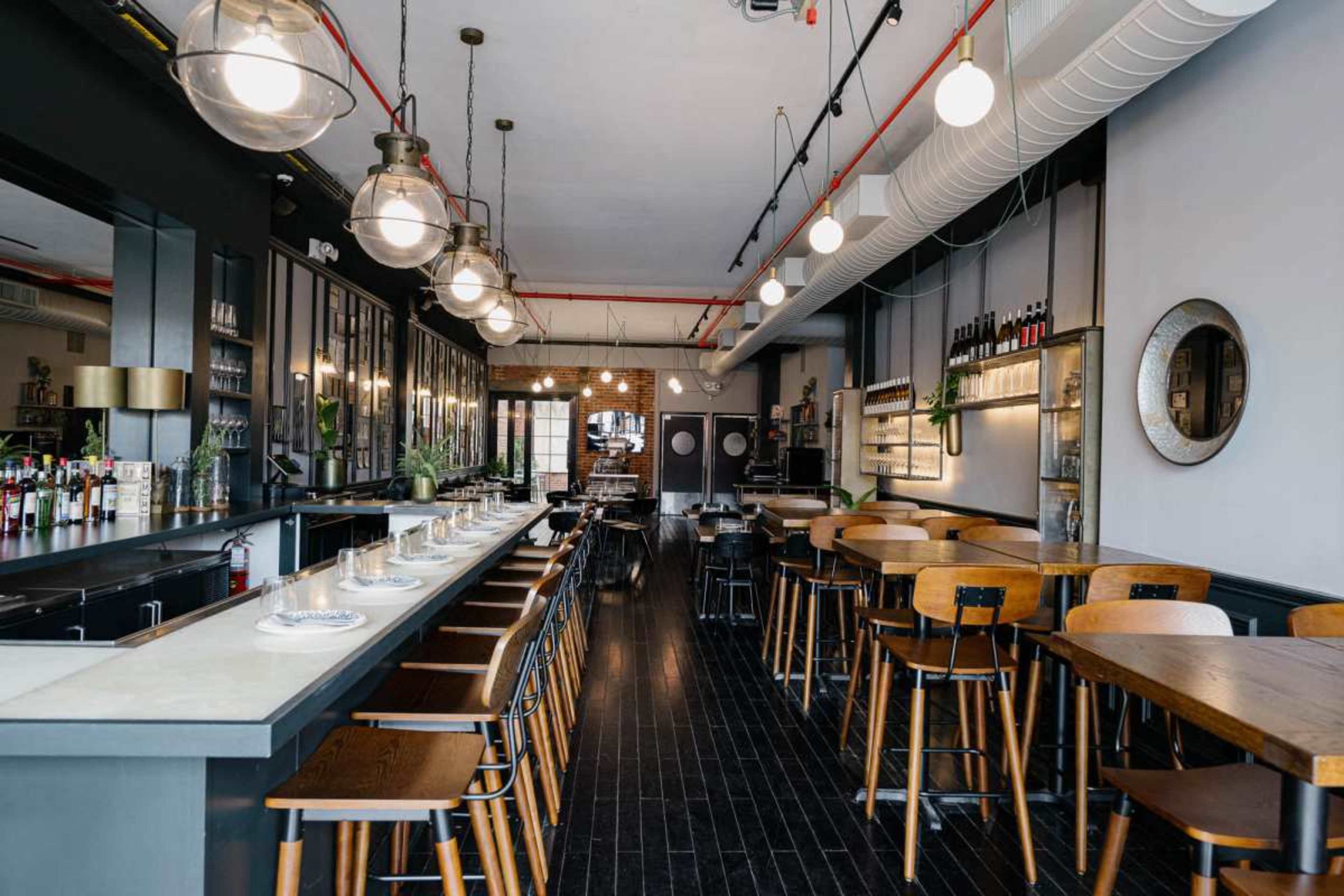 The image shows an interior view of a modern restaurant featuring a long bar with stools, wooden tables, and pendant lighting.