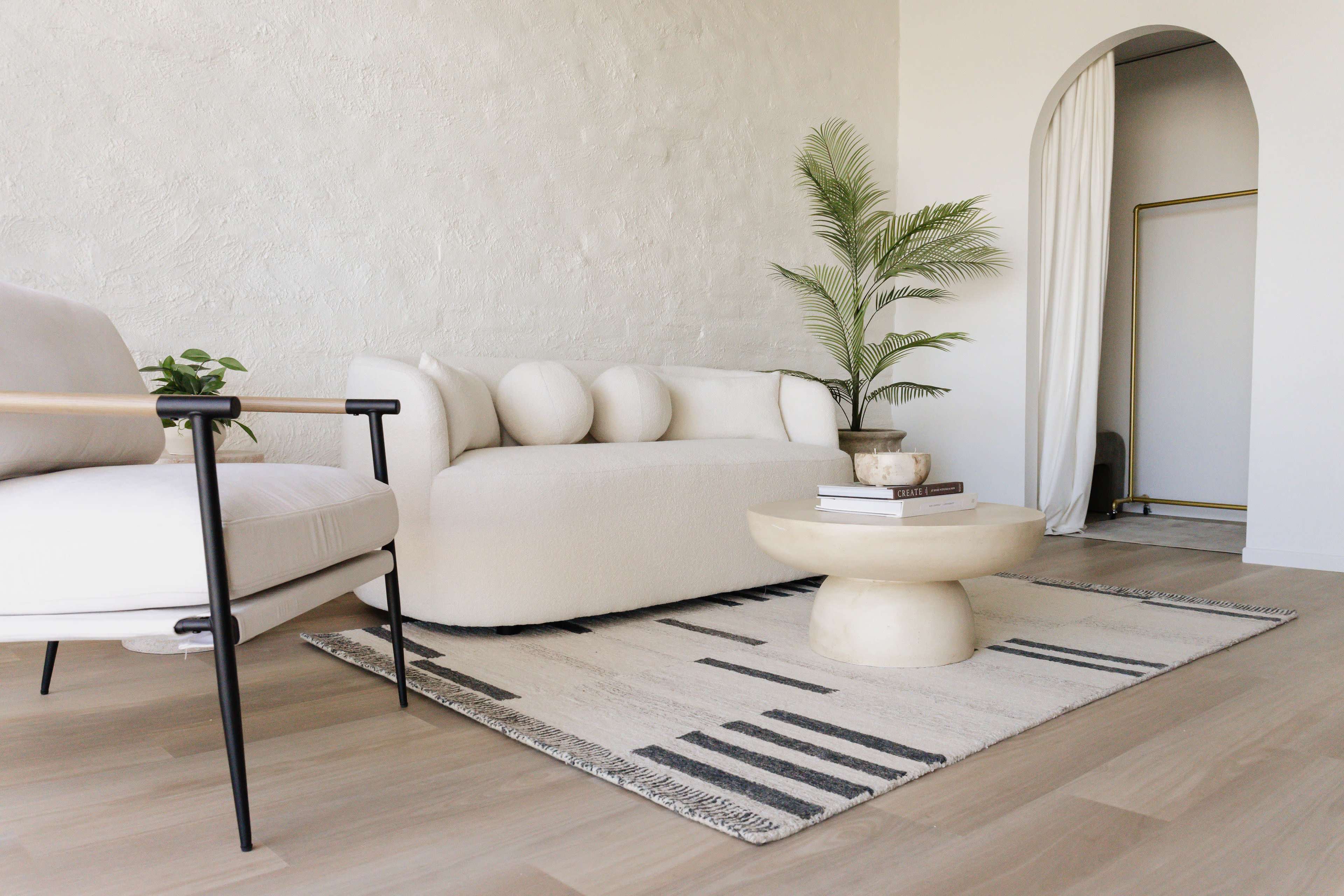 The image shows a minimalist living room featuring a light-colored sofa, a round coffee table, and a potted plant on a textured rug.