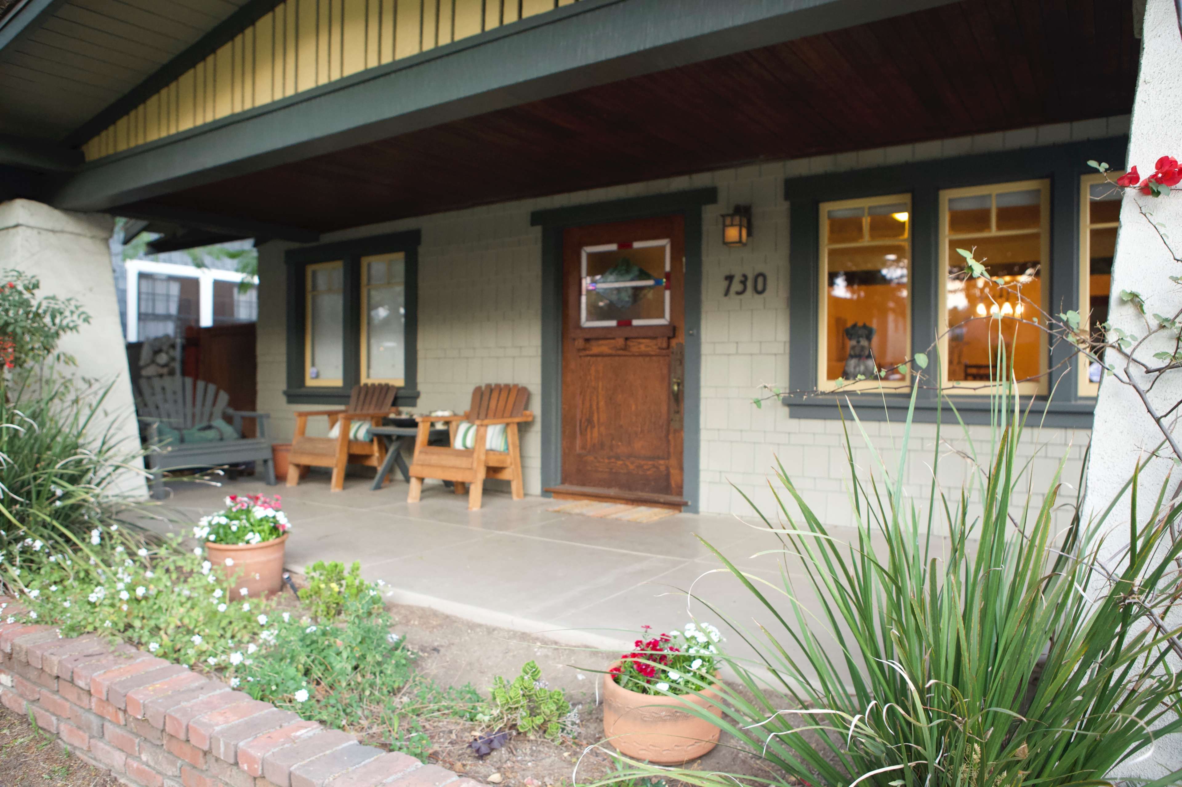A porch with wooden chairs and a front door is surrounded by potted plants and greenery.