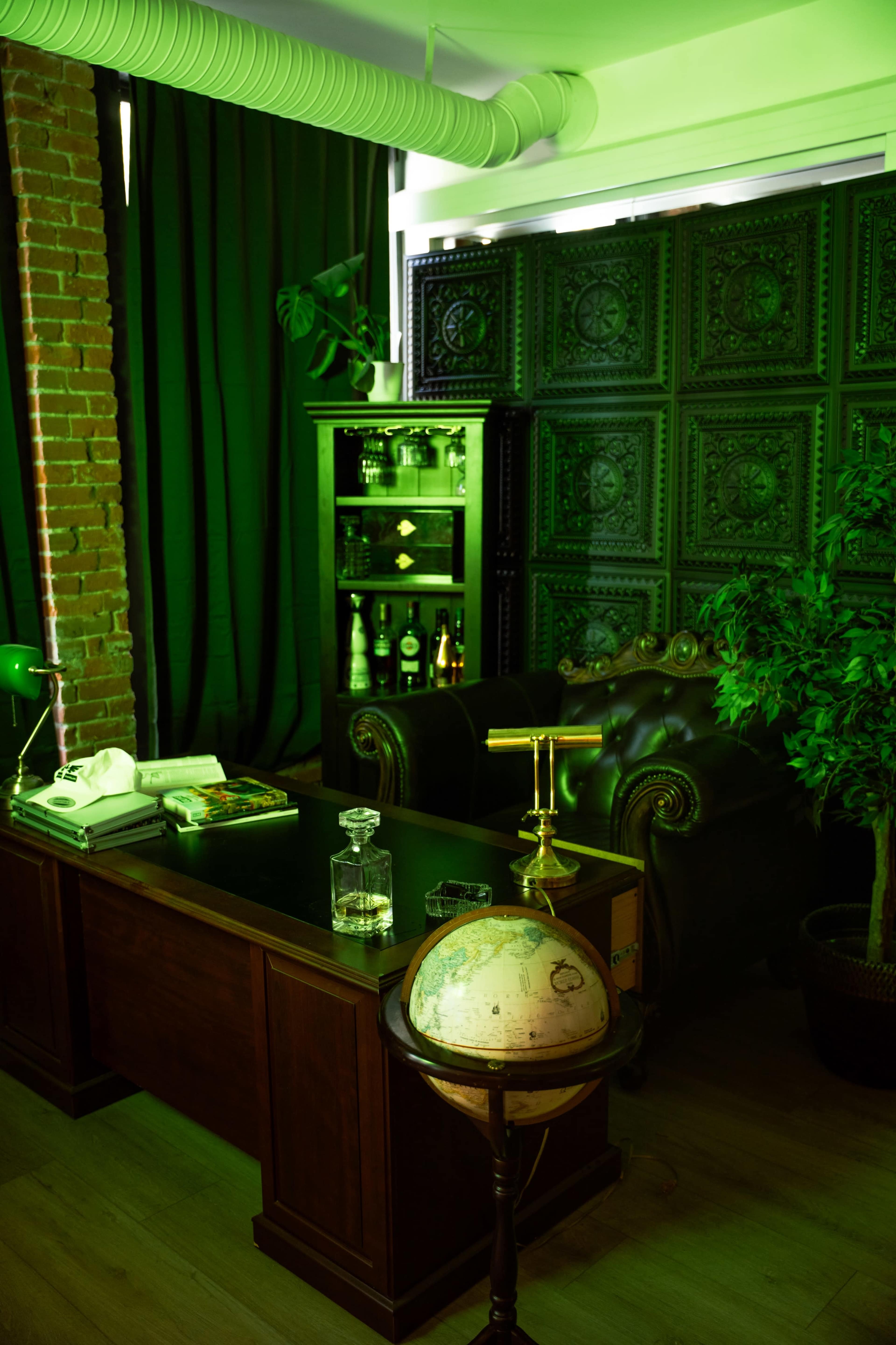 The image shows a stylish office space featuring a dark wooden desk, a leather armchair, a globe on a stand, and shelves with bottles against a backdrop of green lighting and decorative wall panels.
