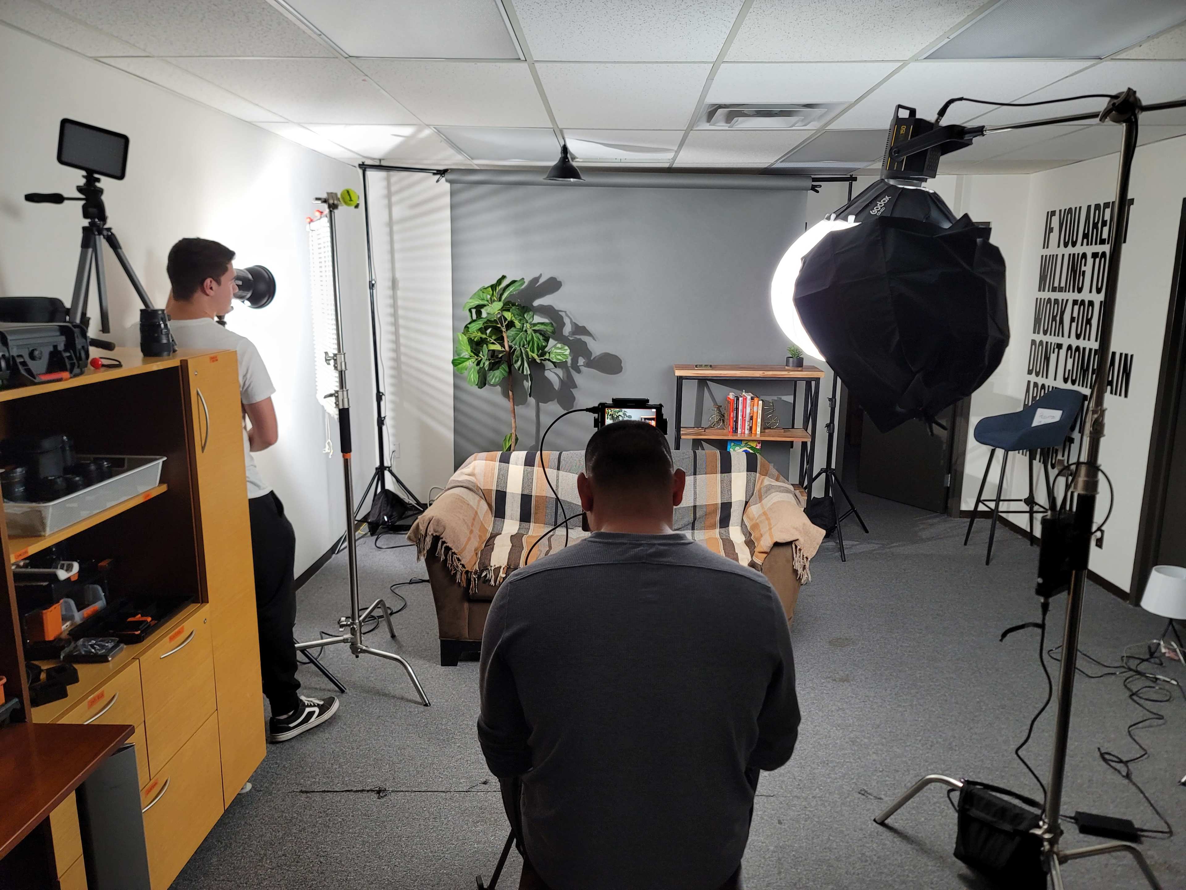A person sits on a couch in a well-lit studio setting with equipment and a backdrop while another person operates a camera.