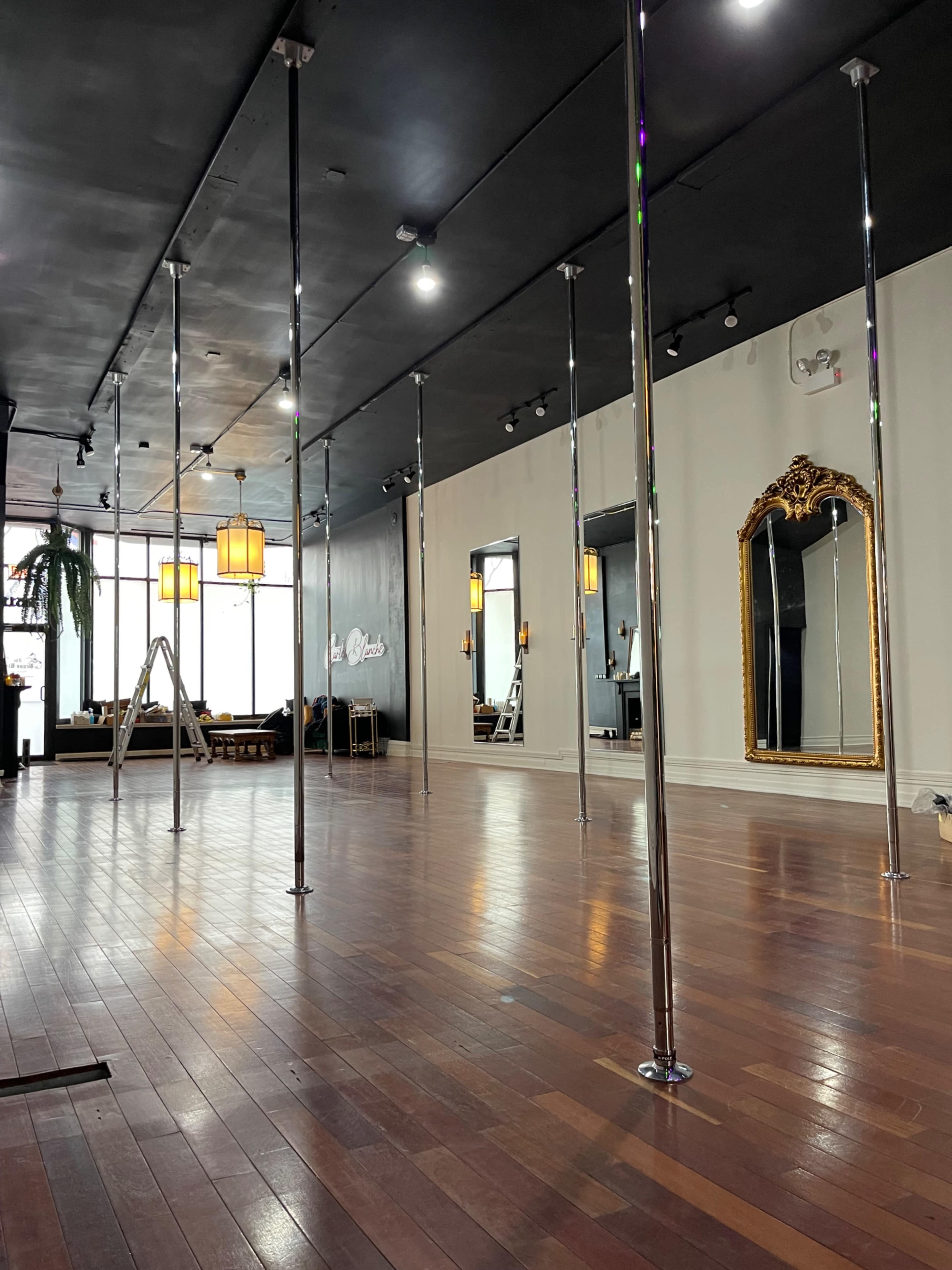 An indoor studio with polished wooden floors, several vertical poles, and mirrors along the walls, illuminated by overhead lights.