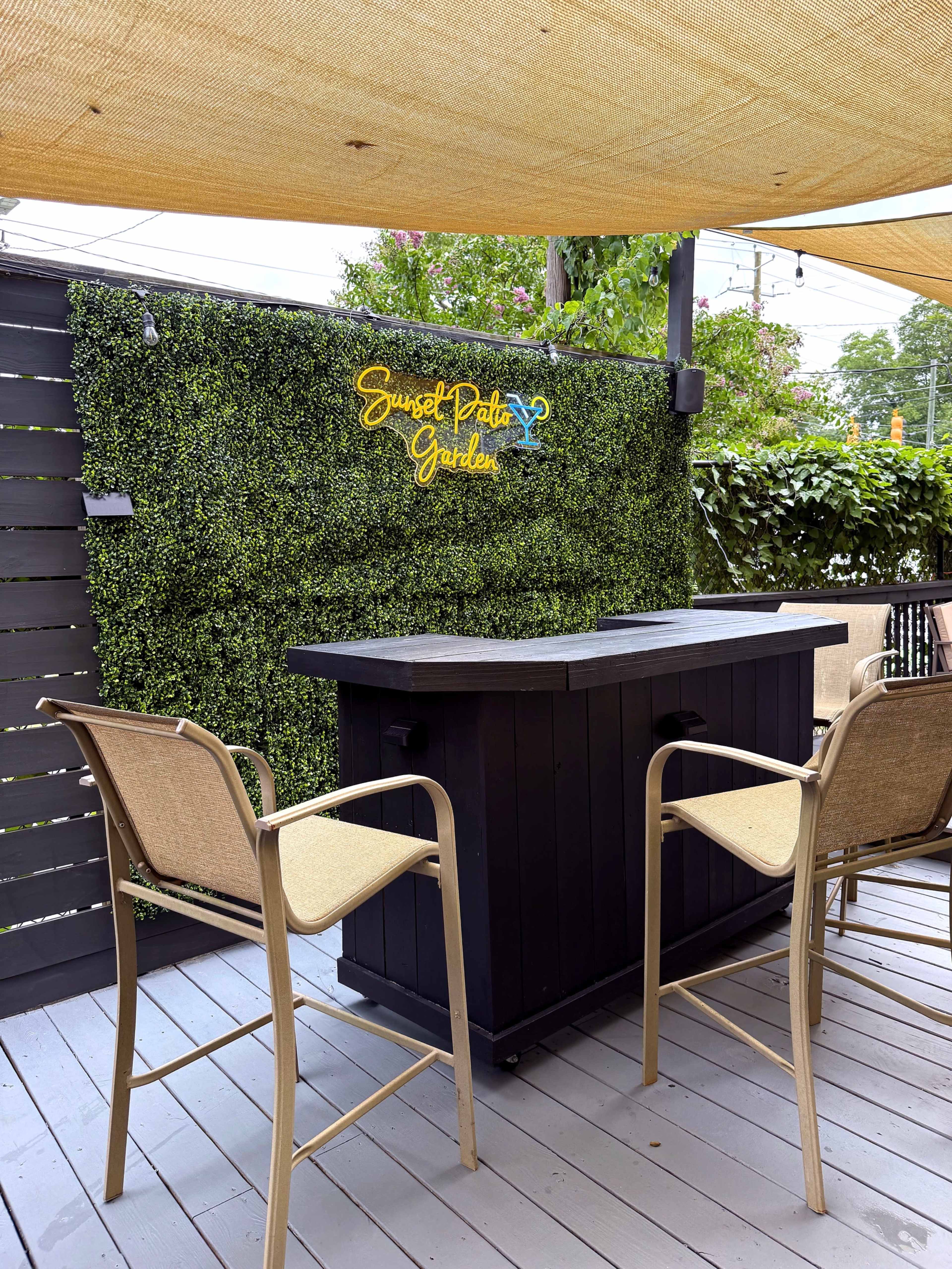 A black outdoor bar is set against a green wall covered in artificial foliage, with the sign "Sunset Patio Garden" illuminated above it.