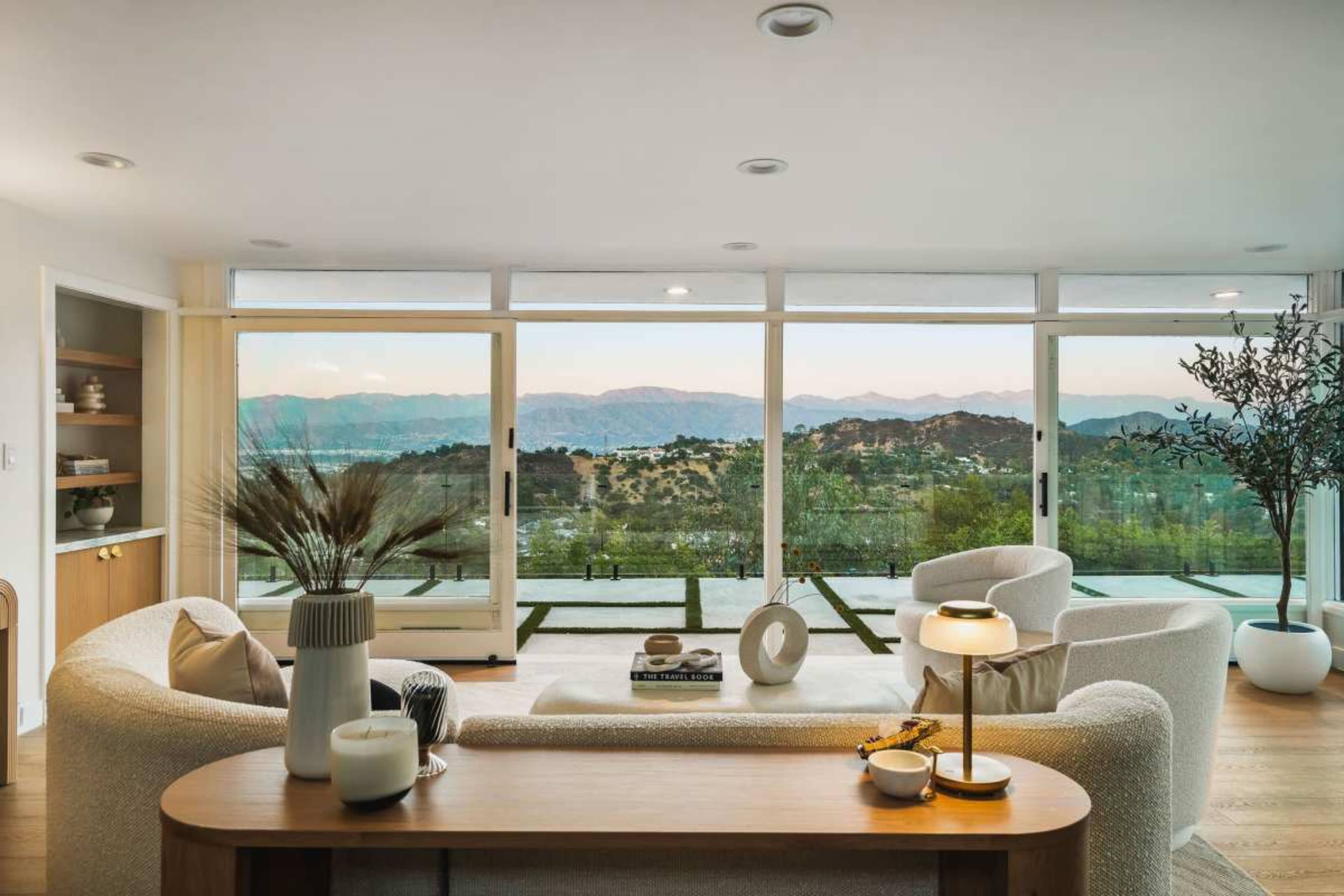 A modern living room features a curved sofa, a wooden console table, and large windows with a view of rolling hills and mountains.