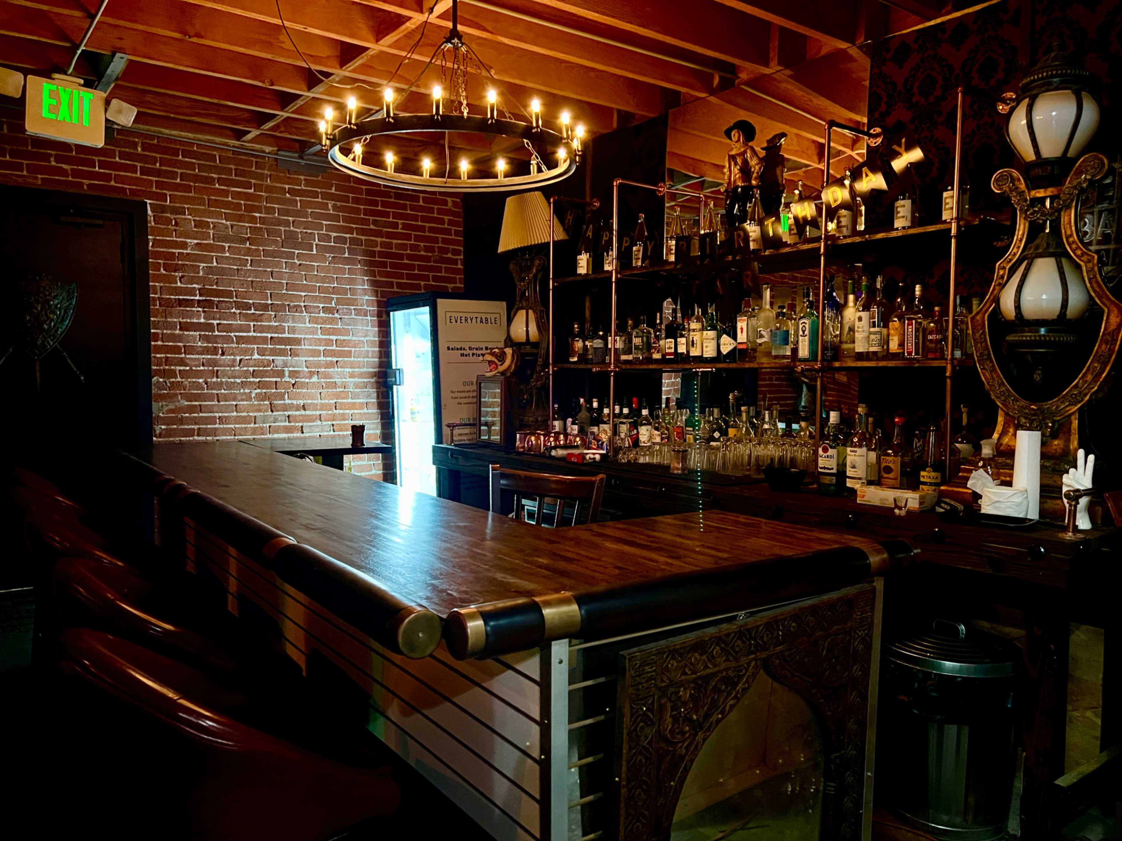 A rustic bar area with a long wooden counter, a chandelier overhead, and shelves stocked with various bottles of liquor against a brick wall.