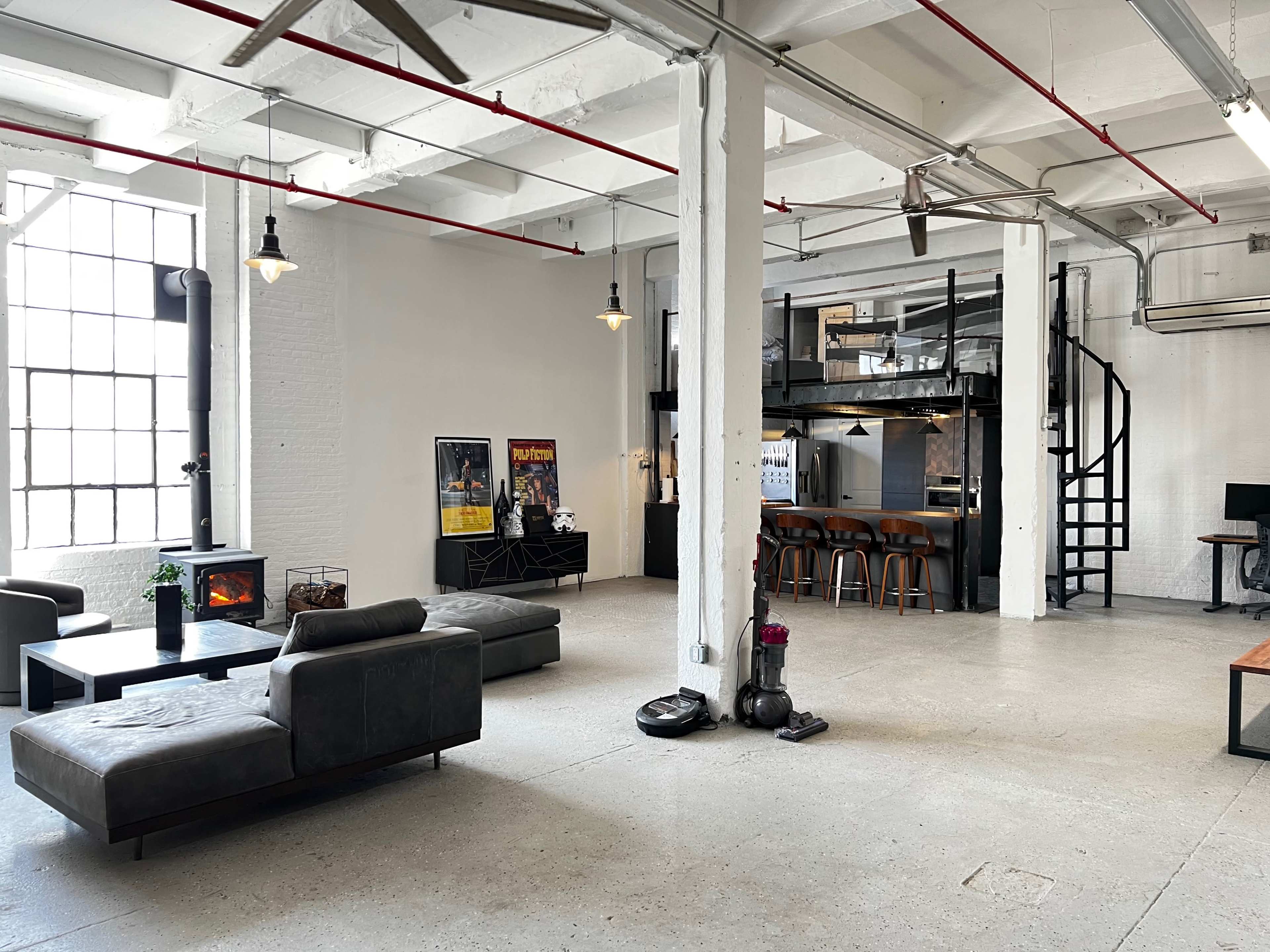 A spacious, industrial-style loft with a living area, a staircase leading to an upper level, and a kitchen featuring barstool seating.