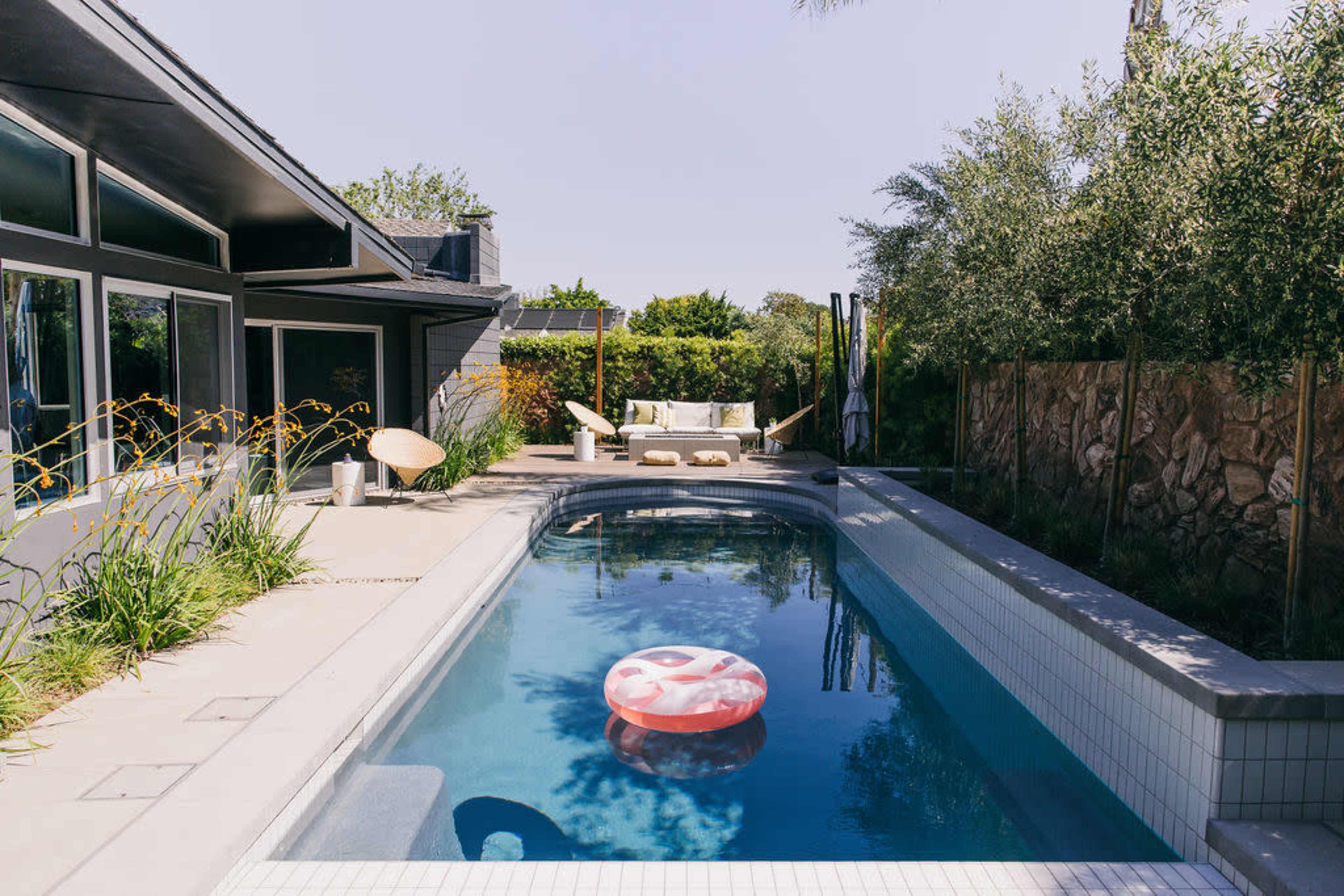 A swimming pool with a red inflatable float is surrounded by a patio and greenery near a modern house.