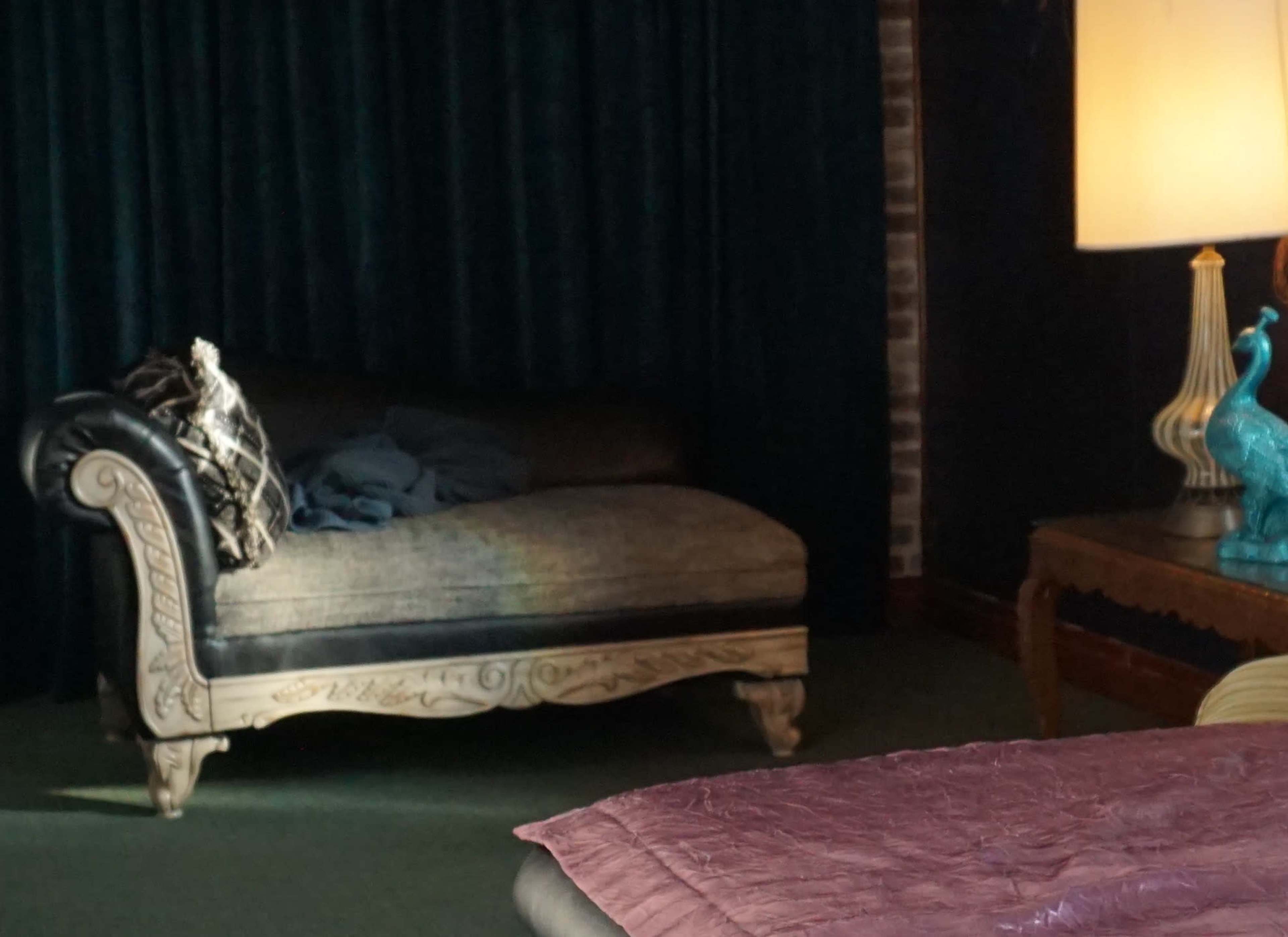 A vintage chaise lounge with ornate legs sits next to a lamp and a decorative peacock figurine in a room with dark curtains.