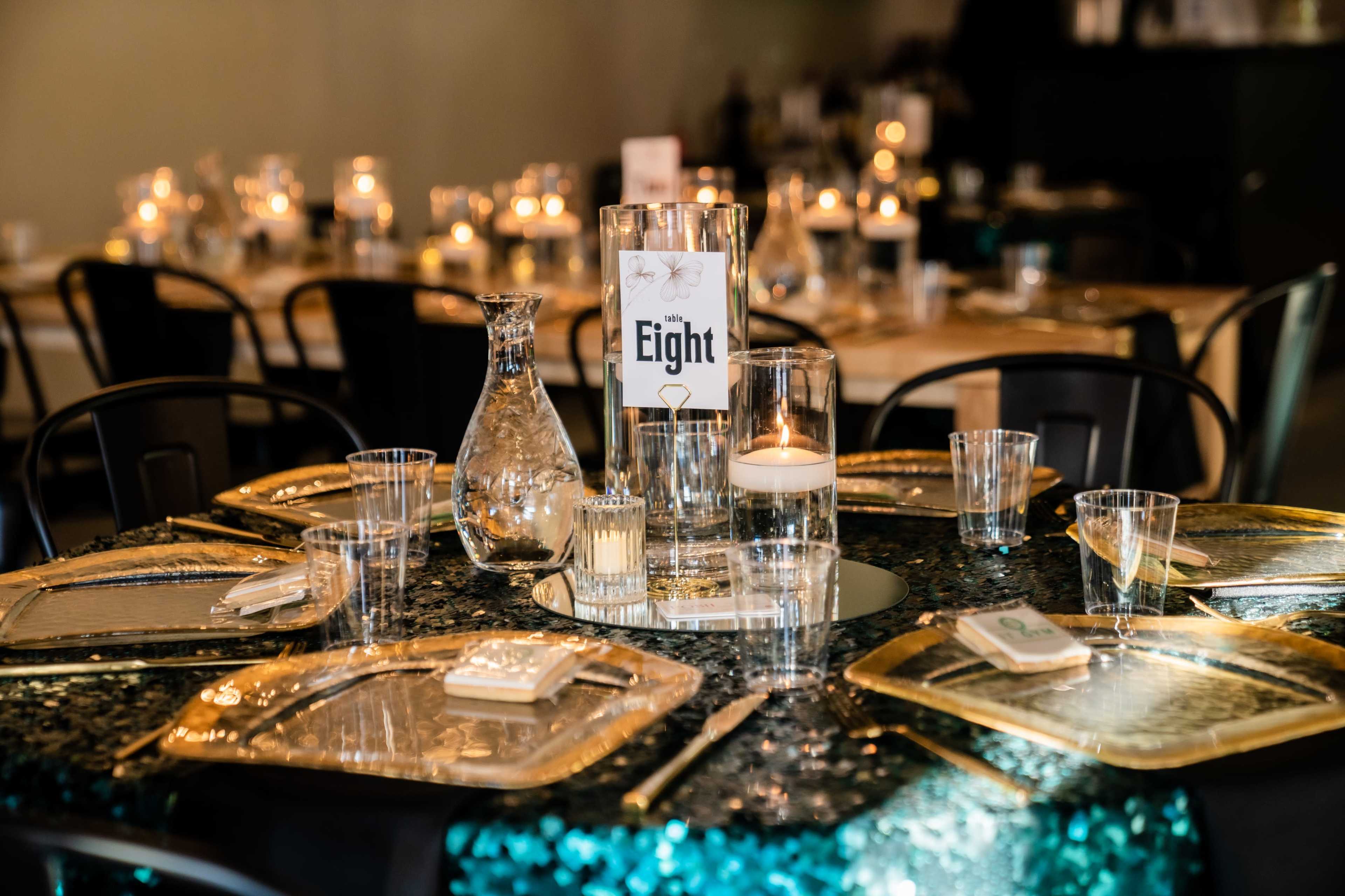 A round table is elegantly set with glassware, silver trays, a decorative tablecloth, and a centerpiece displaying the number "Eight."