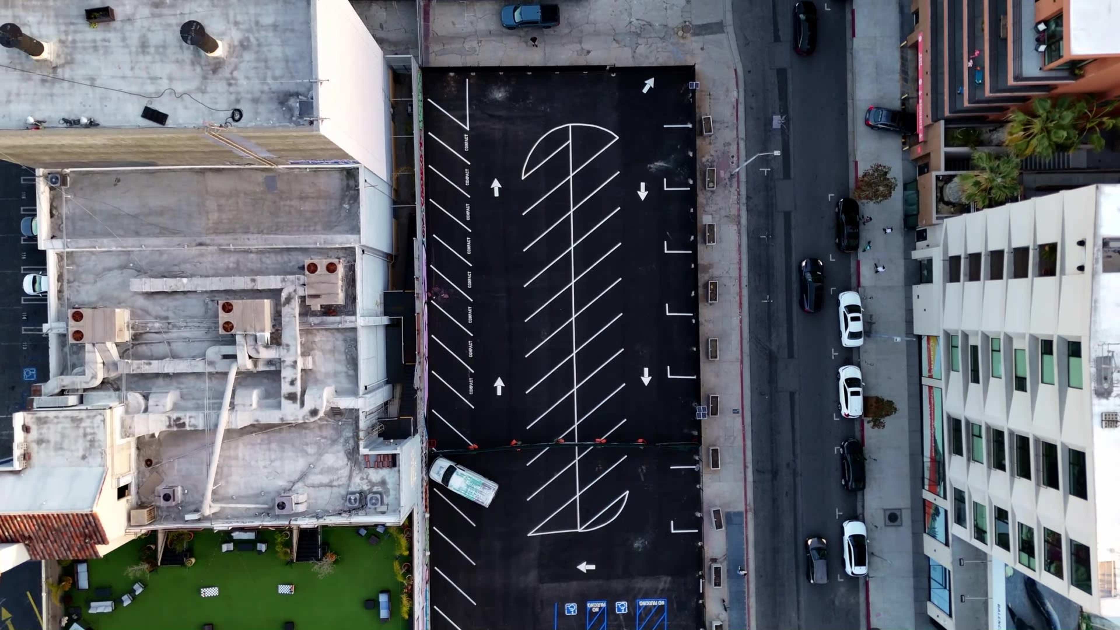 The image shows an overhead view of a parking lot with distinct white lines marking parking spaces, adjacent to a street lined with parked cars.