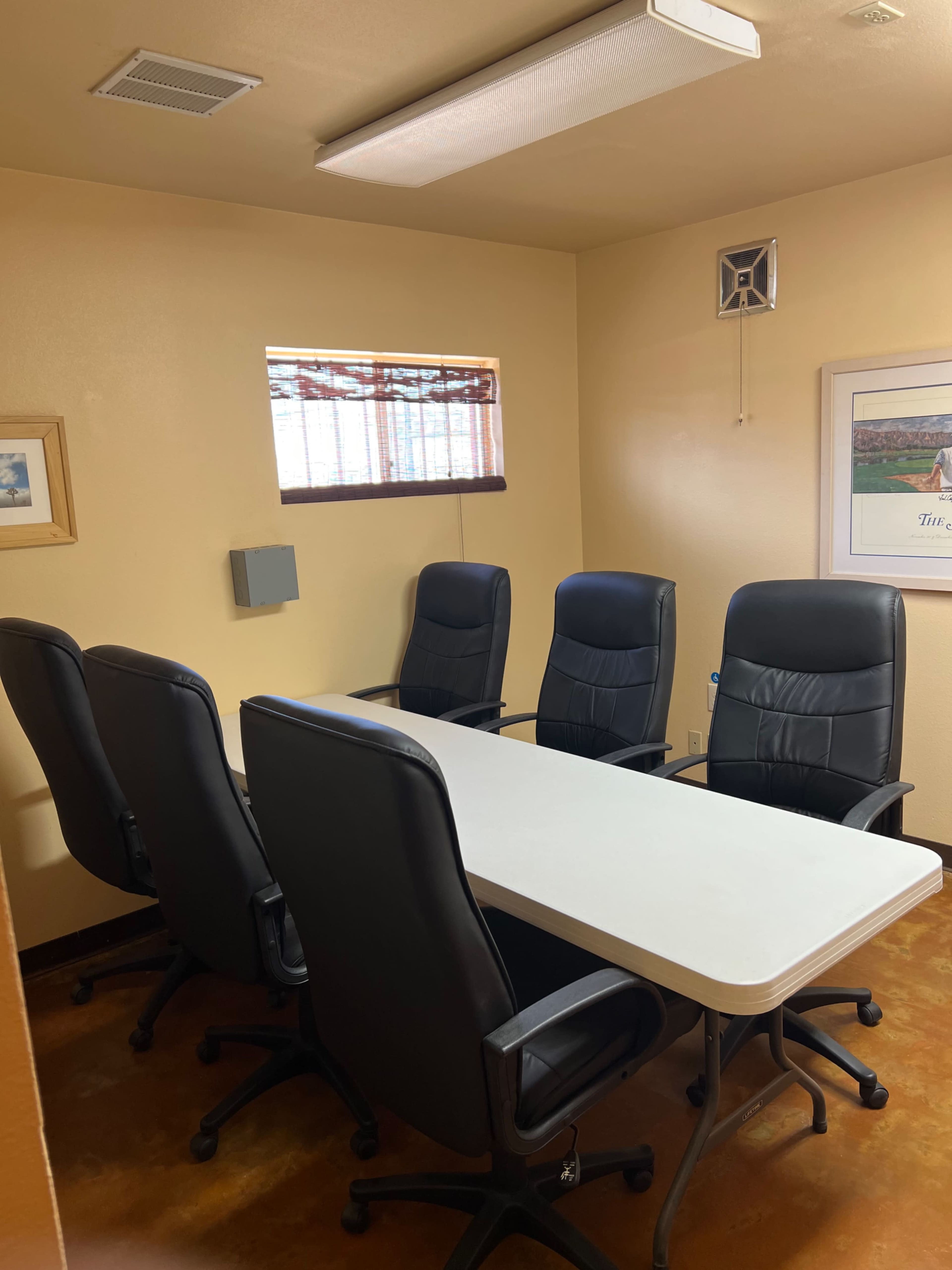 The image shows a small conference room with a long table and five black chairs arranged around it.