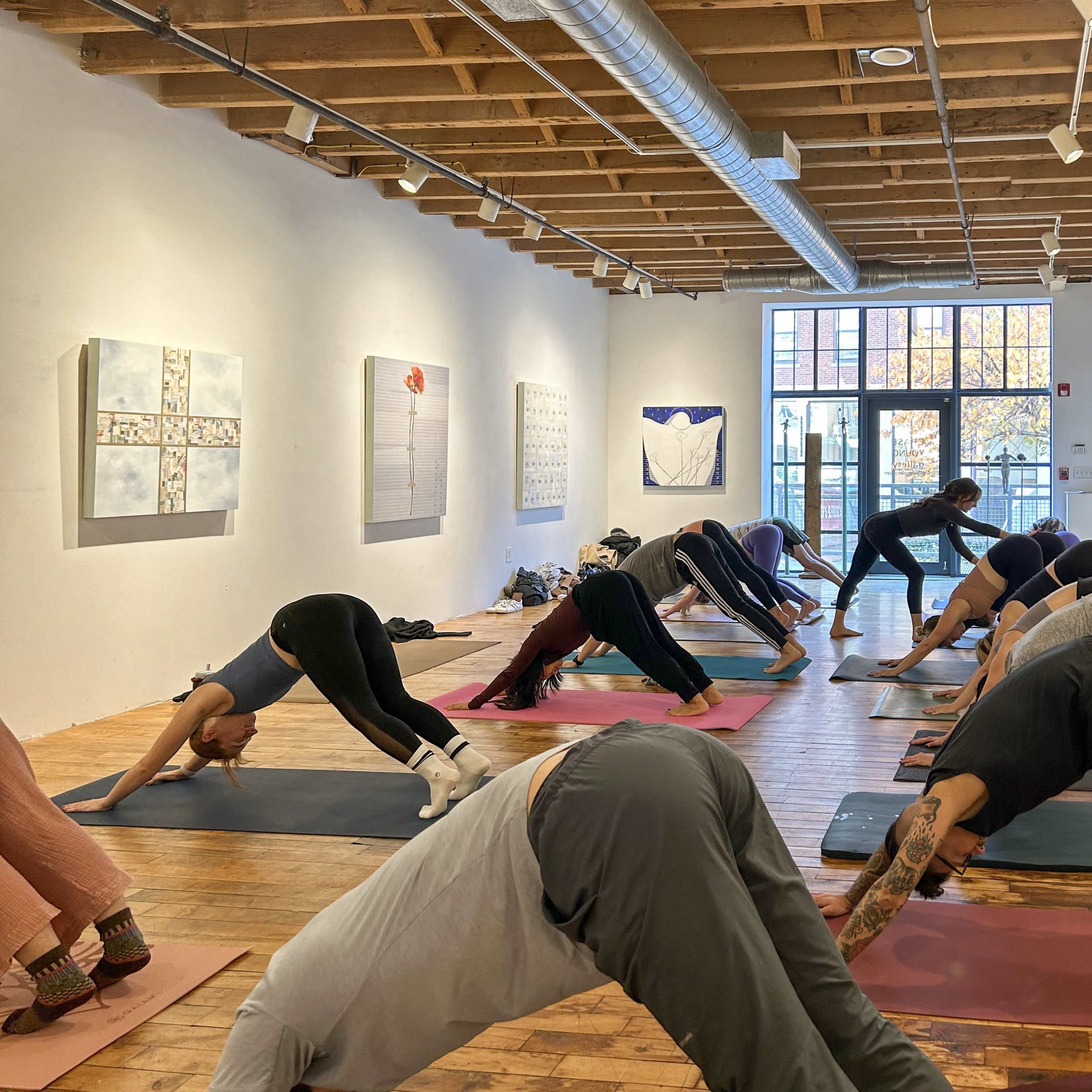 A group of people in a yoga class performs downward-facing dog poses on mats in a spacious studio with artwork on the walls.