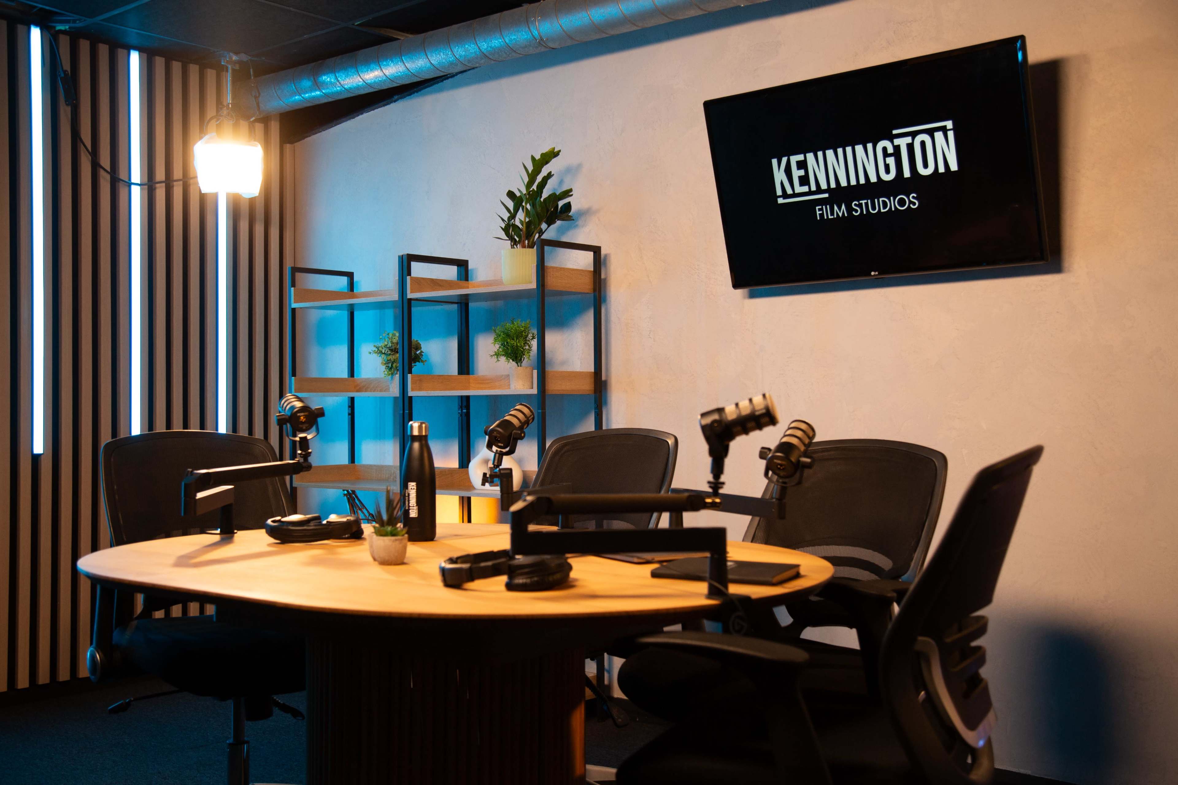 The image shows a podcast studio with a round wooden table, four black chairs, and two microphones set up, alongside a shelf with small plants and a TV displaying the logo "KENNINGTON FILM STUDIOS."