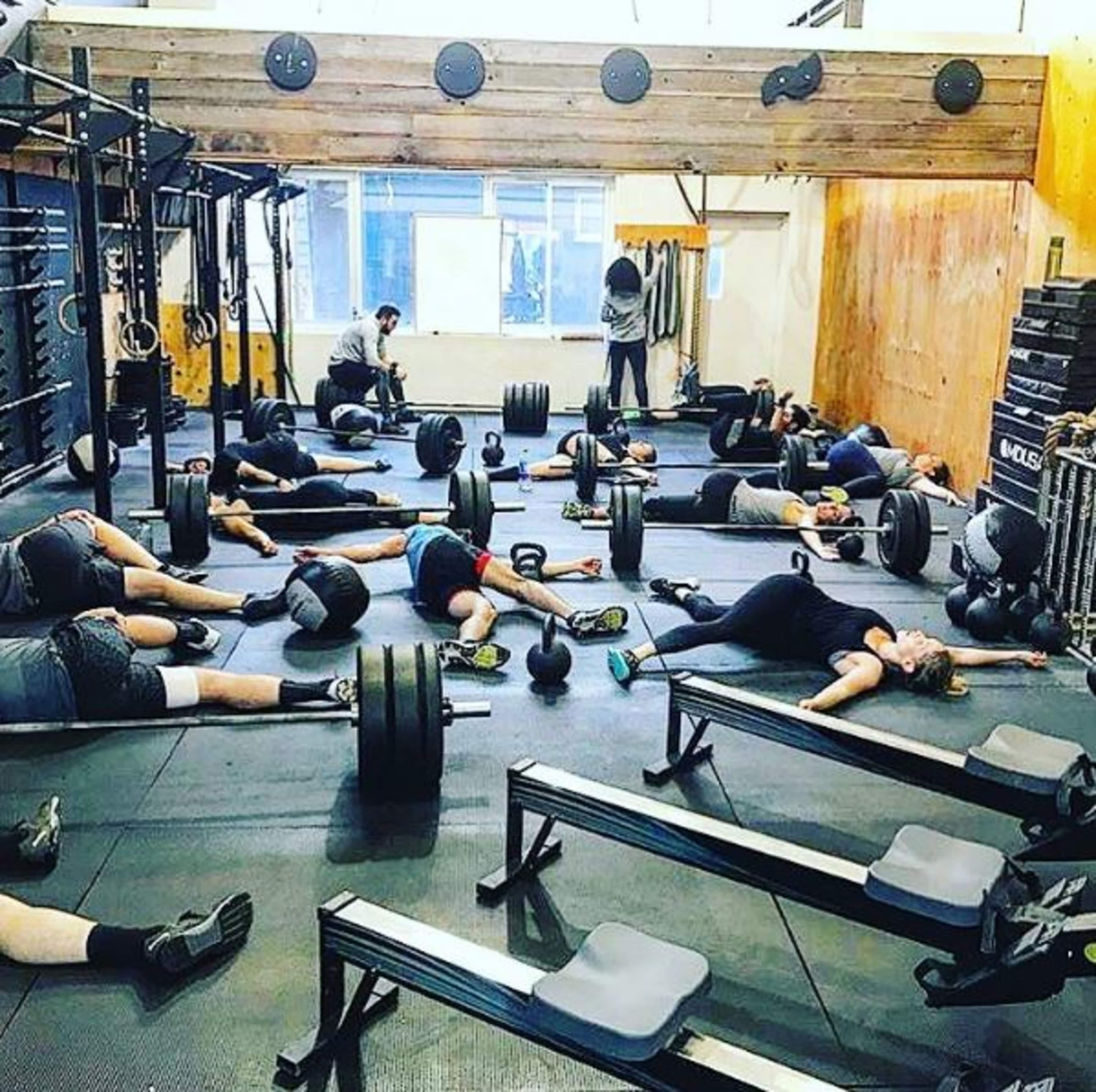 A fitness gym is filled with people lying on the floor among weightlifting equipment and benches after a workout session.