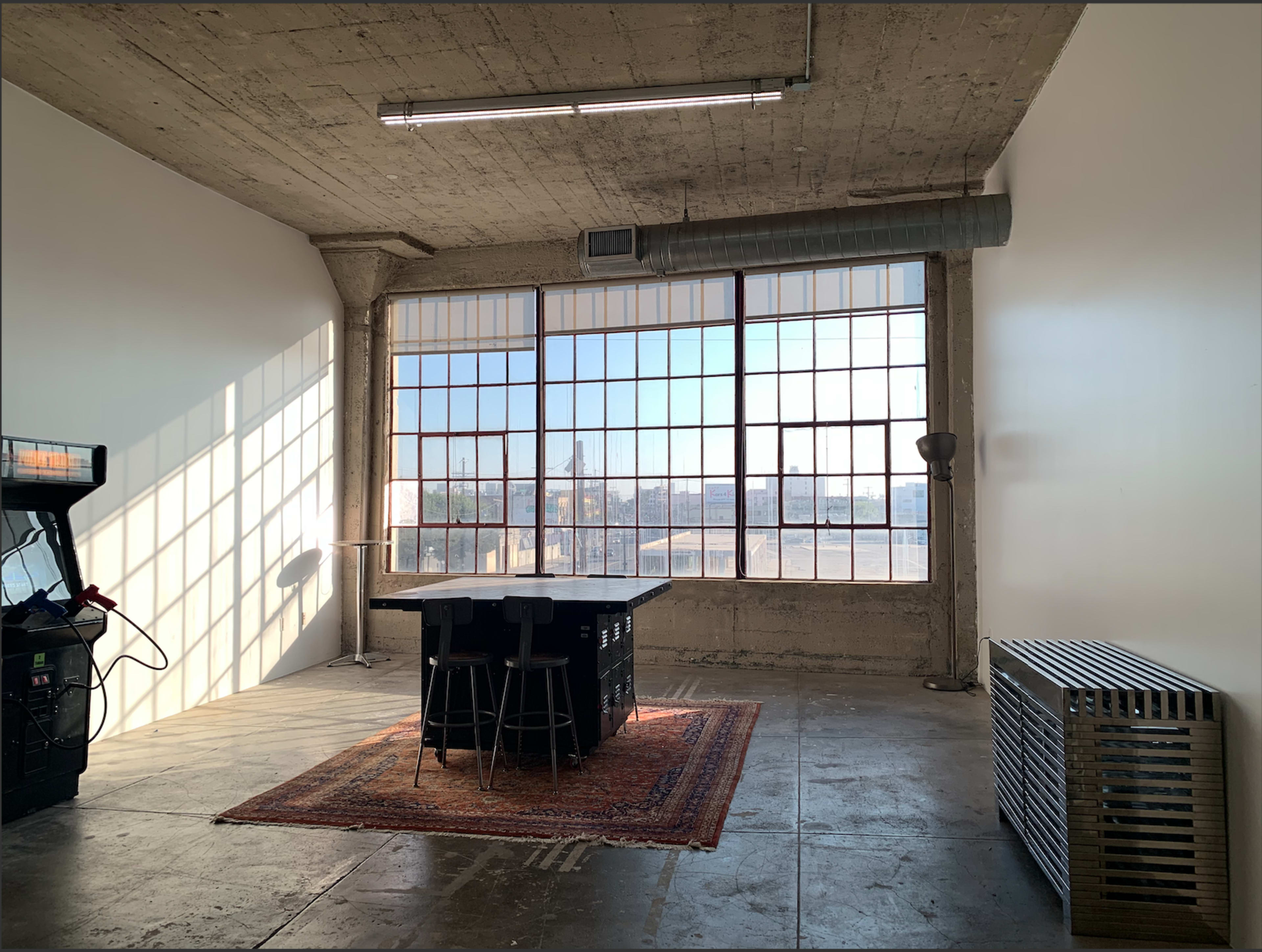 A spacious room with large windows, concrete walls, a rug, a table with stools, and an arcade machine in the corner.