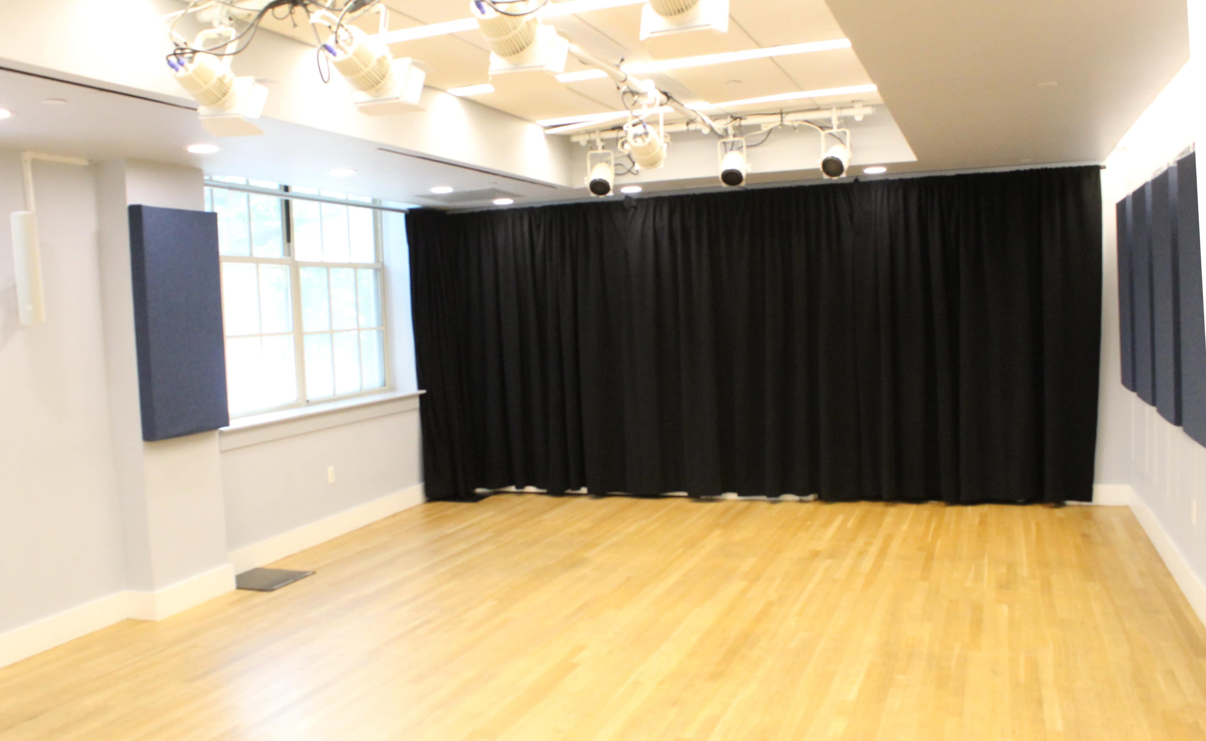 The image shows a spacious, well-lit room with wooden flooring, a window on one side, and black curtains covering a wall.