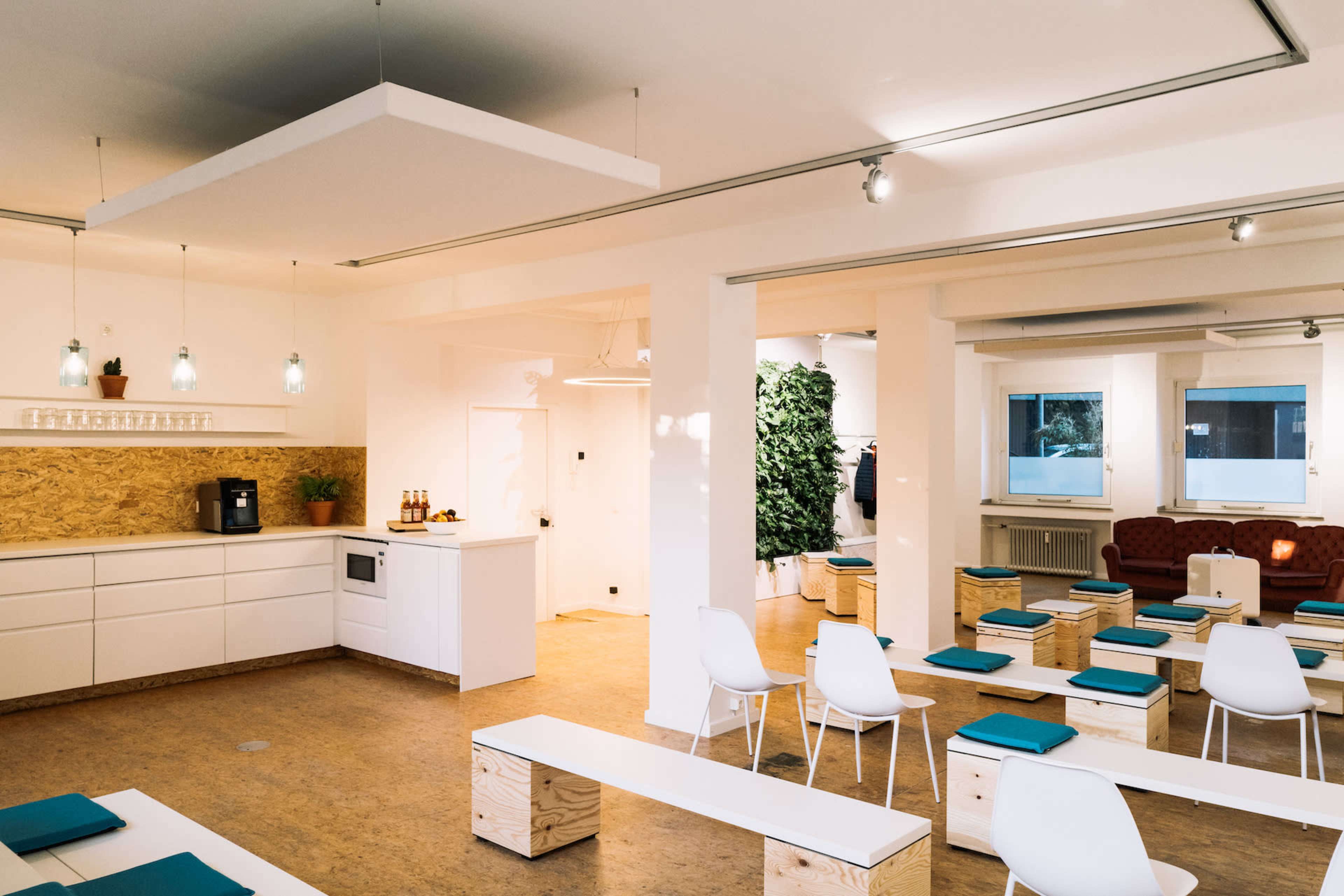 The image depicts a spacious, minimalist room with white walls, wooden flooring, and seating arranged for an event, featuring a kitchenette and plants in the background.