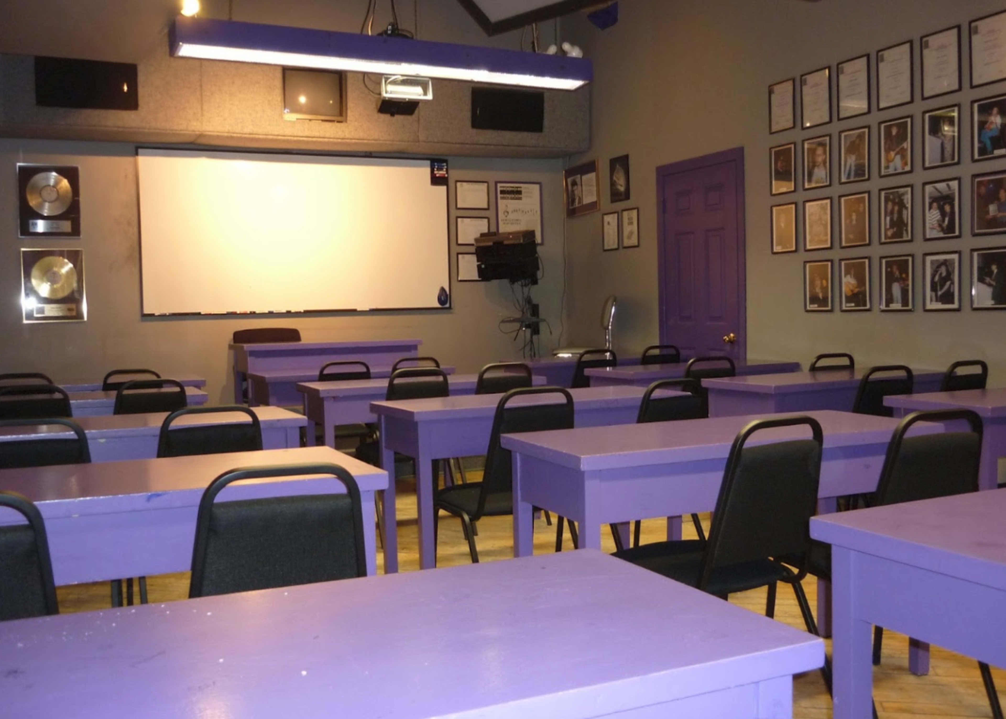 The classroom features purple tables and chairs, with a large white screen at the front and framed certificates on the walls.