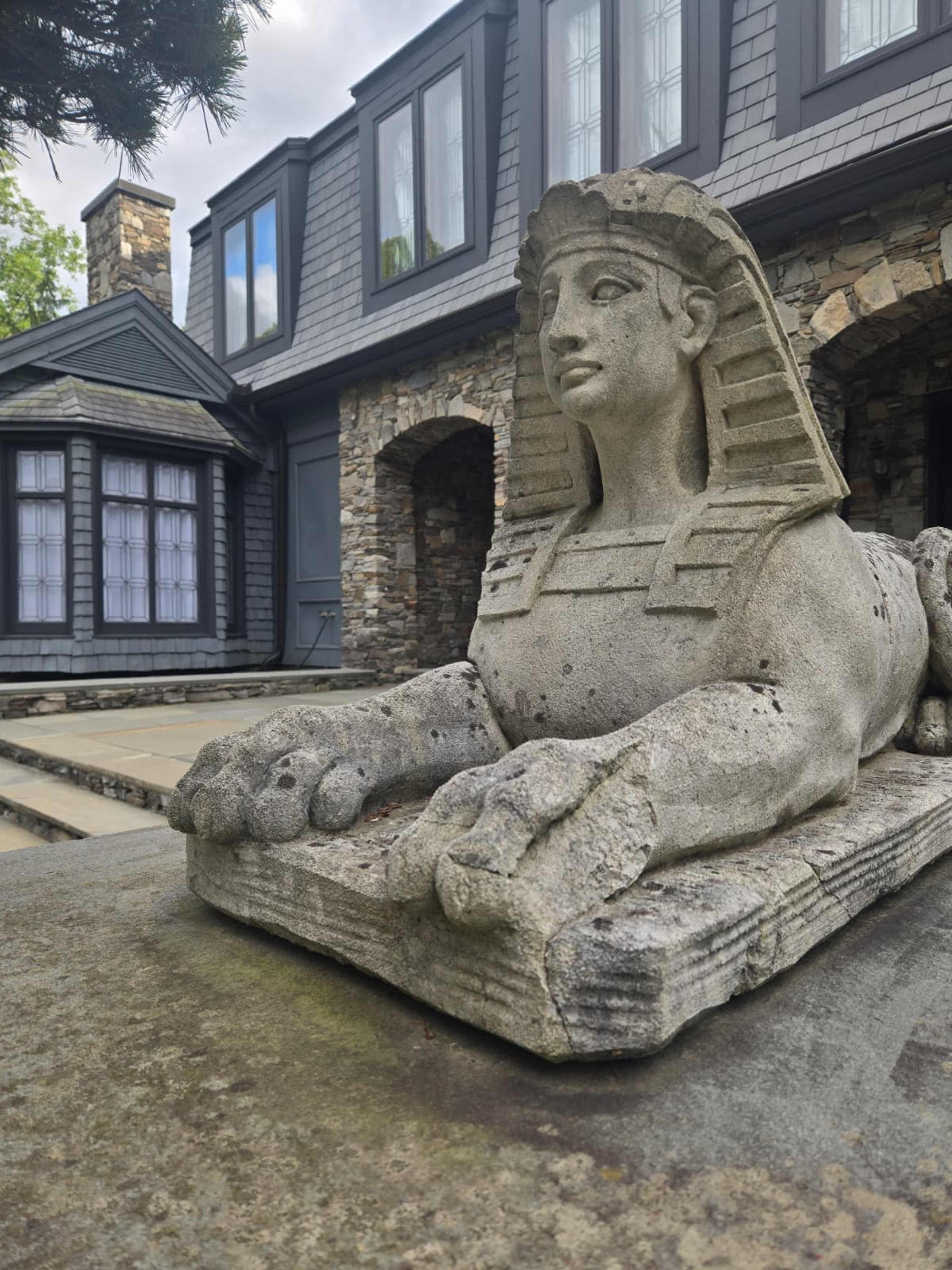 A stone sphinx statue lies in front of a house with a stone facade and gray shingles.