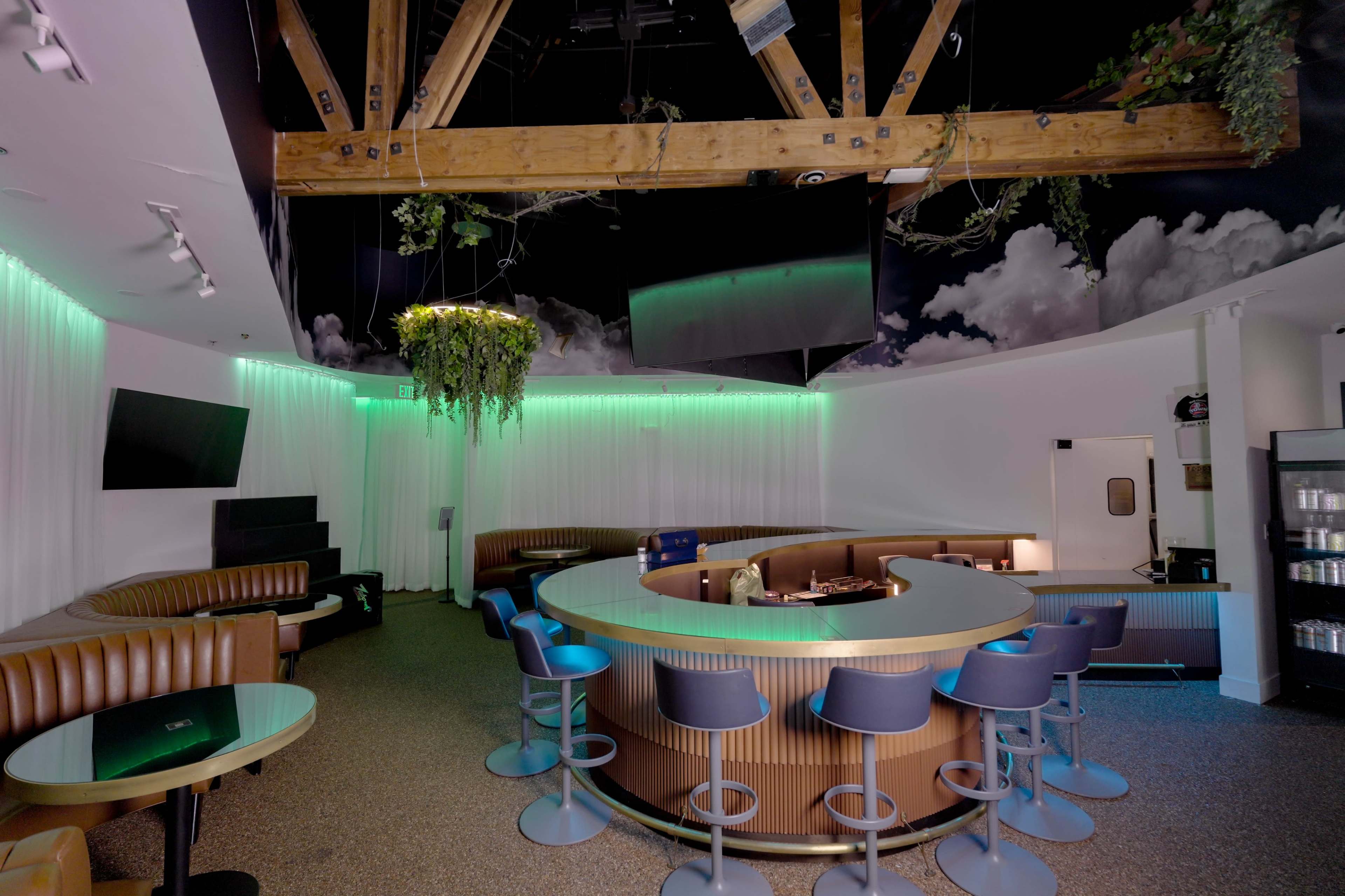 The image shows a modern lounge area with a curved bar, seating along the walls, and greenery suspended from the ceiling under soft lighting.