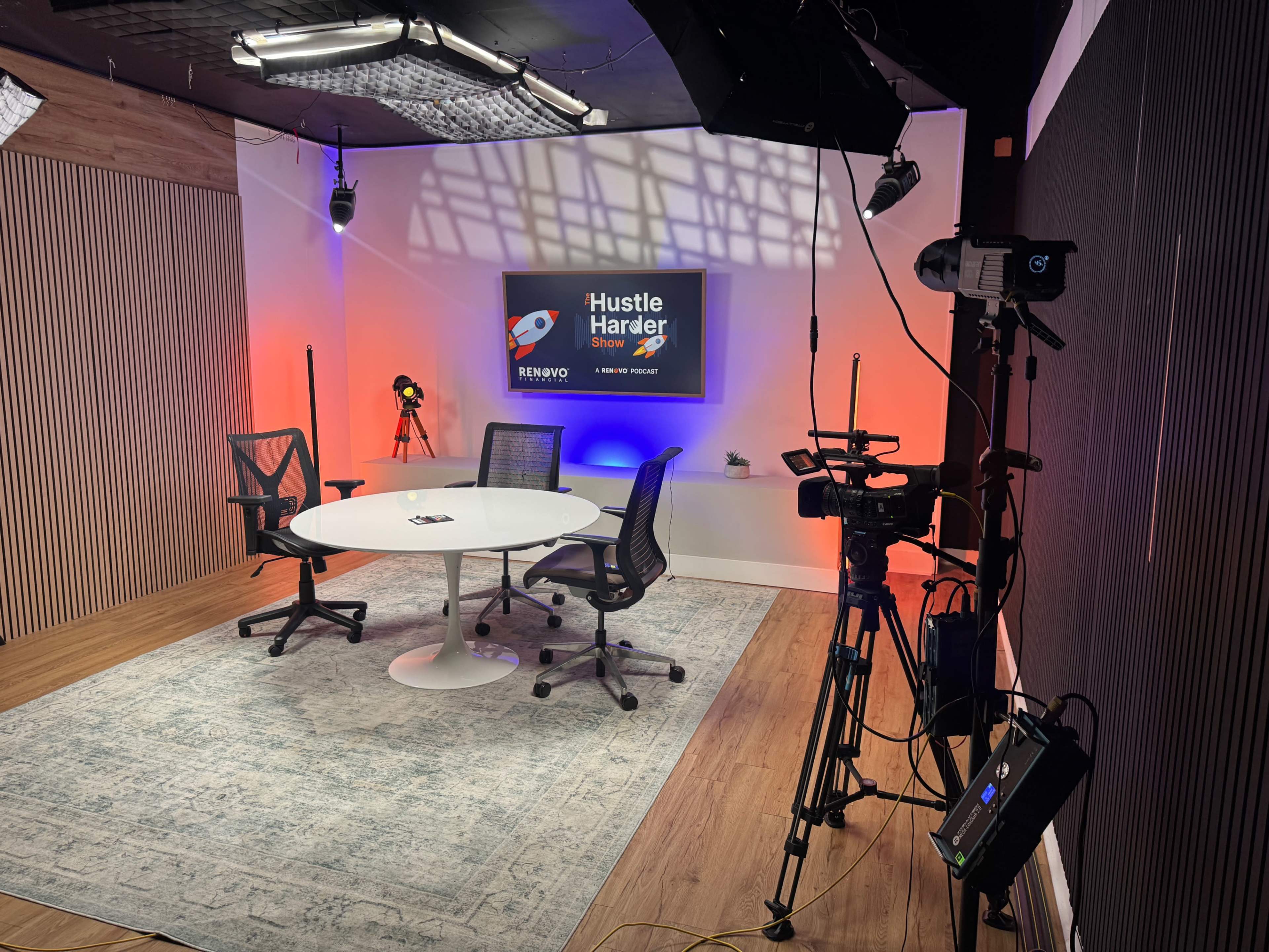The image shows a modern studio setup with a round table and chairs, featuring a screen displaying "Hustle Harder Show," and various lighting and camera equipment around the room.