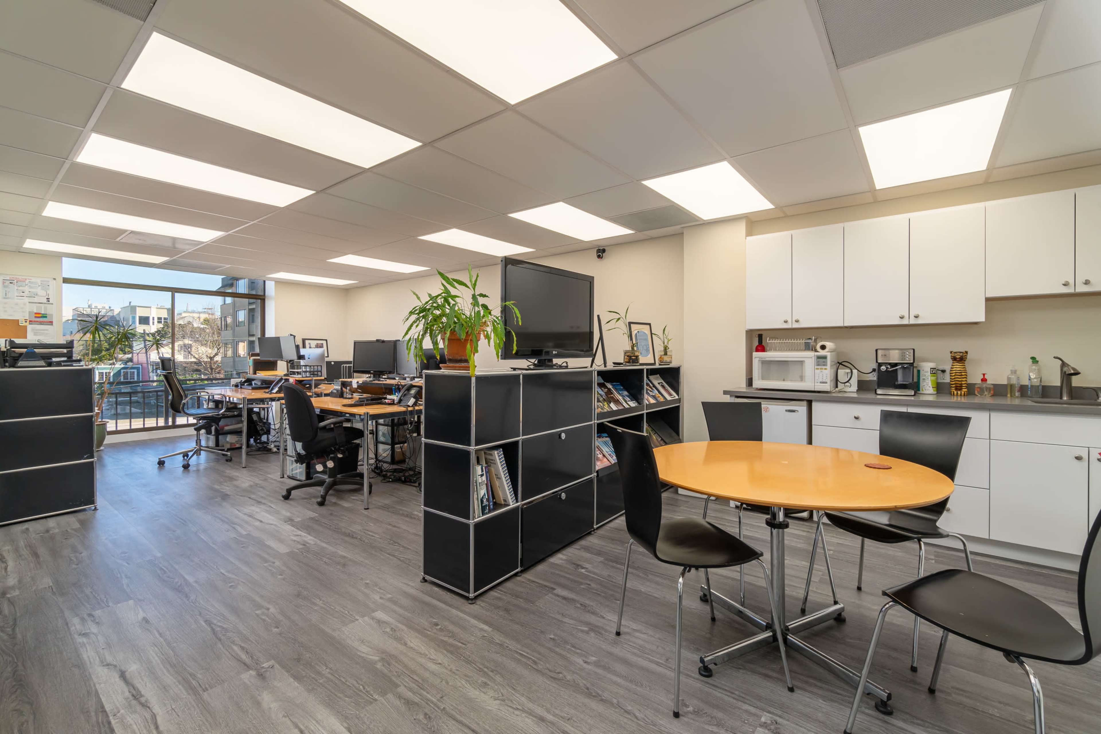 A modern office space featuring workstations, a kitchenette area with appliances, and a seating table surrounded by chairs.