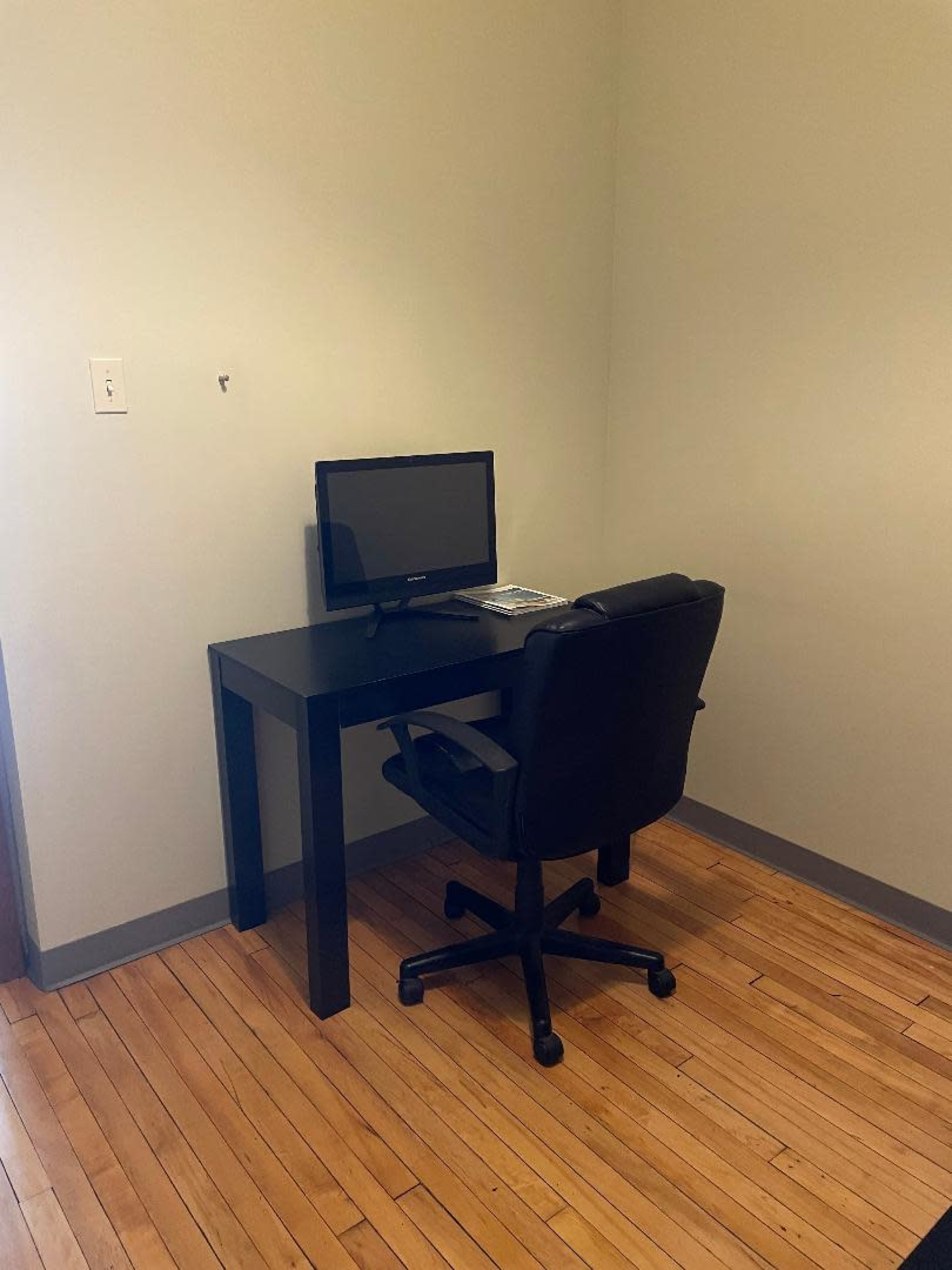 A small corner office setup features a black desk with a computer monitor and a black rolling chair on hardwood flooring.