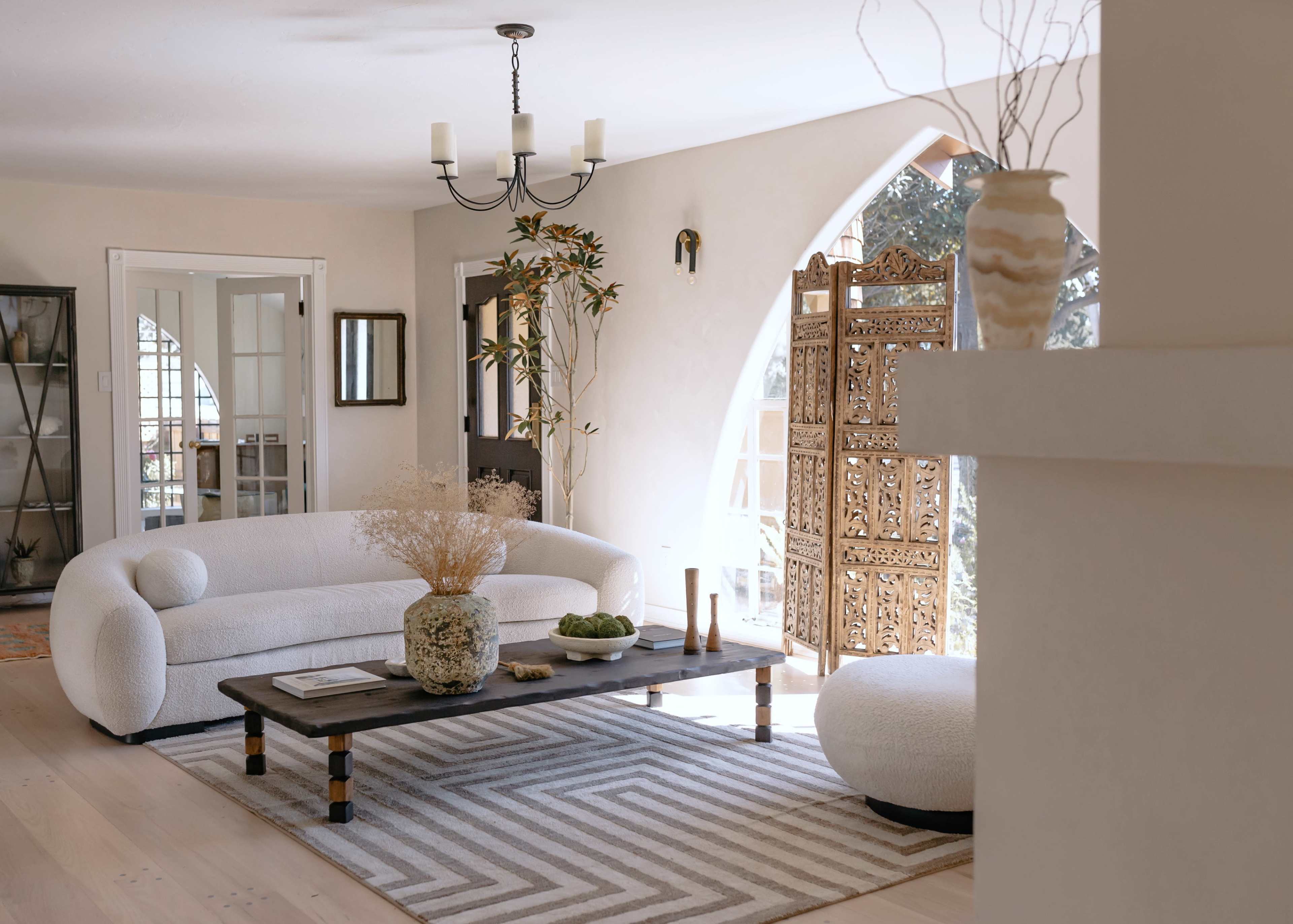 A bright living room with a curved white sofa, a wooden coffee table with decorative items, and an ornate wooden screen near a large window.