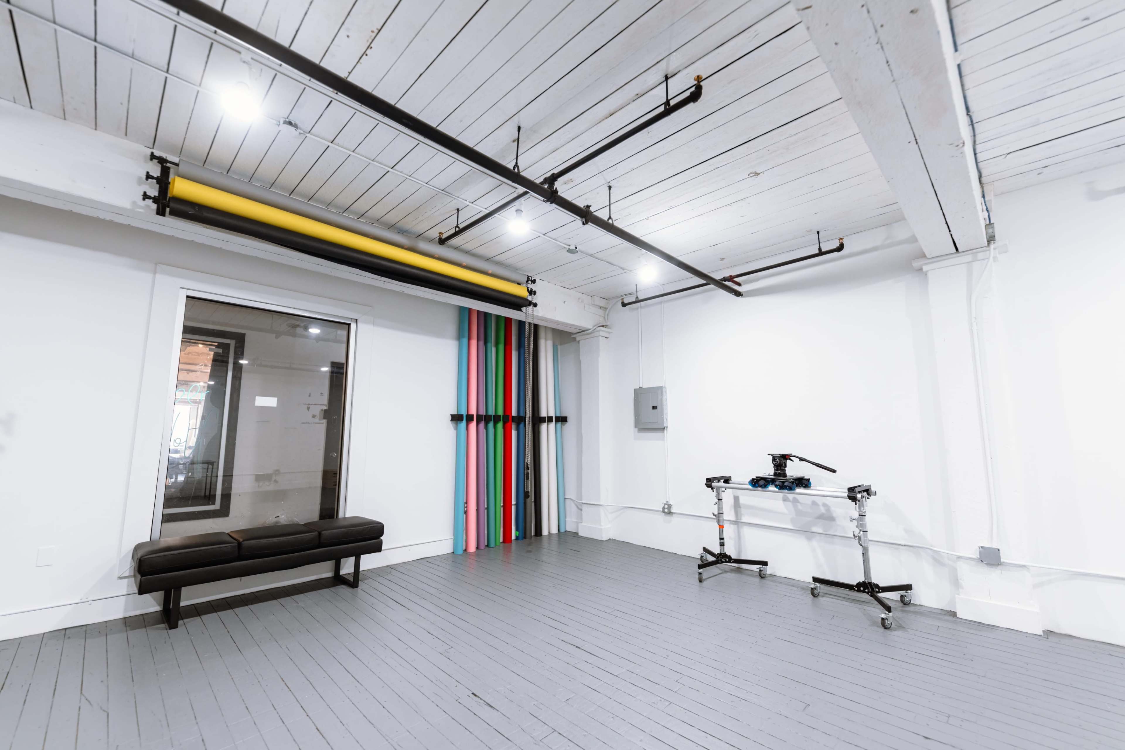 The image shows a minimalist interior space featuring a gray floor, white walls, a black bench, a colorful striped wall, and some overhead pipes and lights.