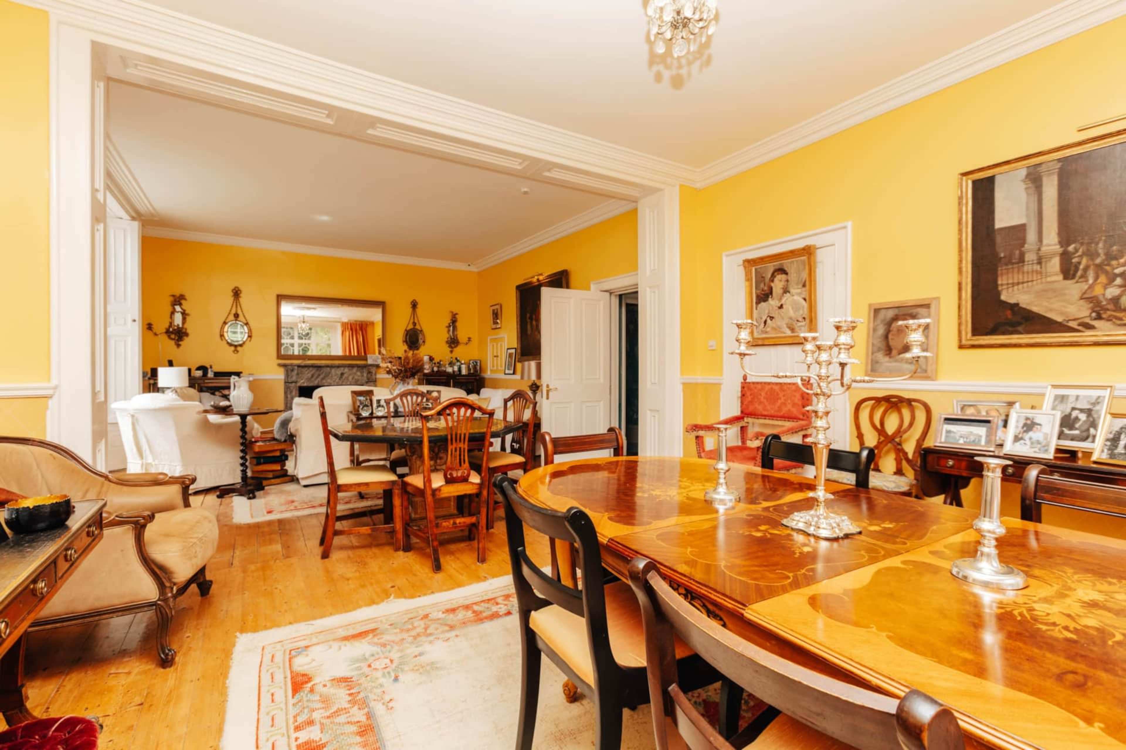 A dining room with a large wooden table, surrounded by chairs, and adorned with various photographs and decorative items against a bright yellow wall.