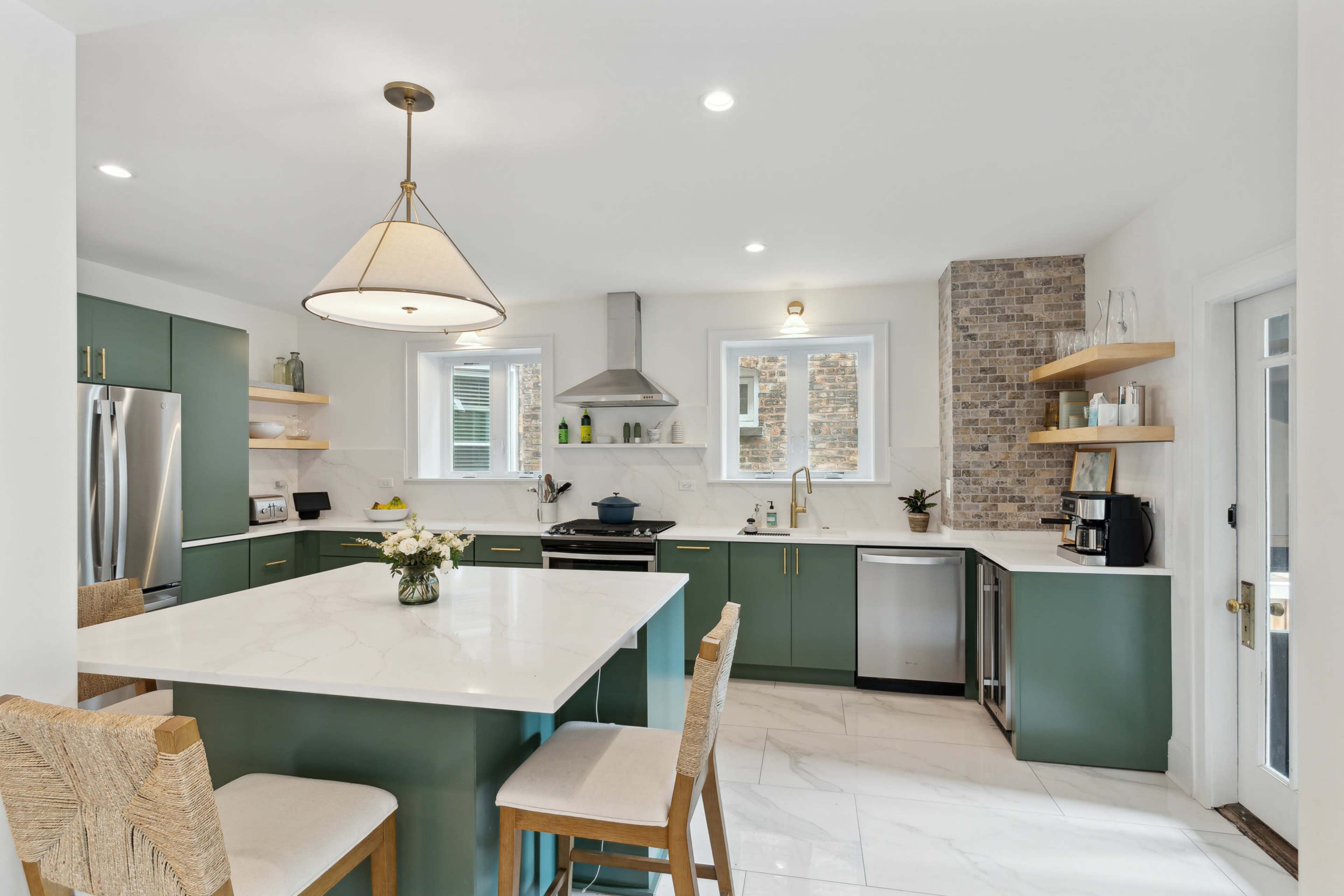 The image shows a modern kitchen featuring green cabinetry, a large marble island with bar stools, and stainless steel appliances.