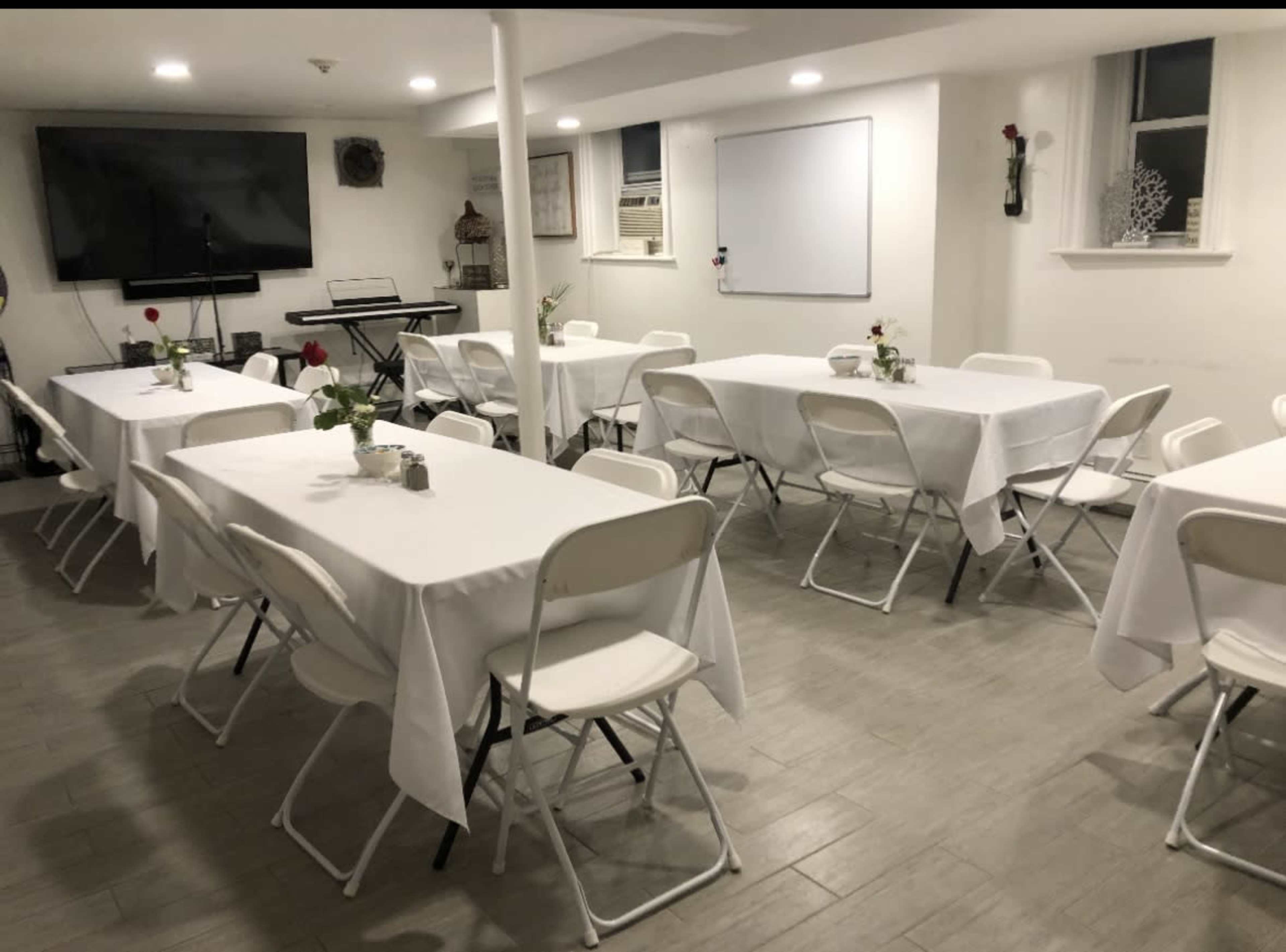 The image shows a spacious room arranged with multiple tables covered in white tablecloths, each set with a small flower arrangement, and equipped with chairs, a large screen, and a keyboard in the background.