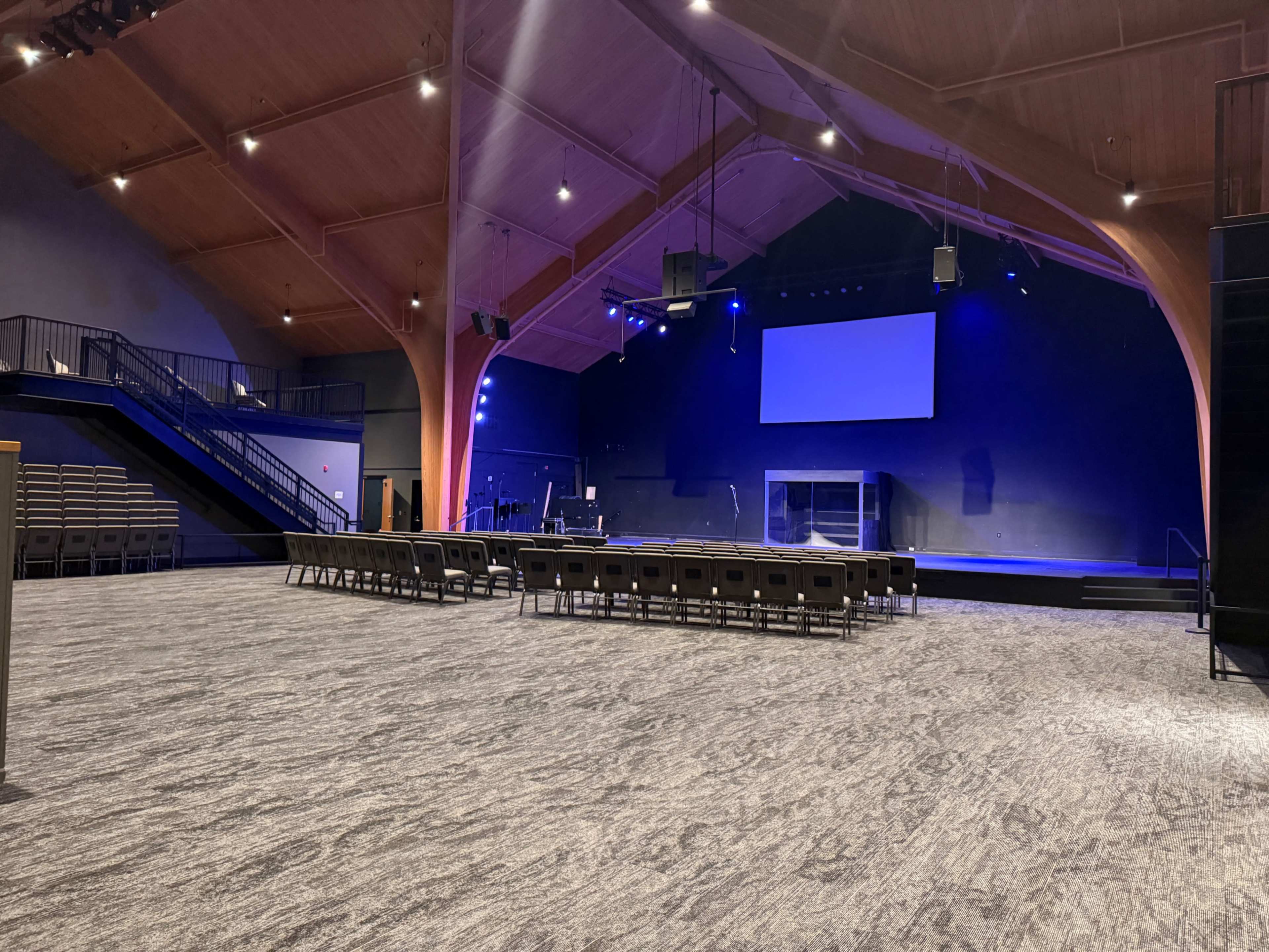 The image shows a spacious auditorium with a wooden ceiling, empty rows of chairs facing a stage equipped with a large screen and stage lighting.