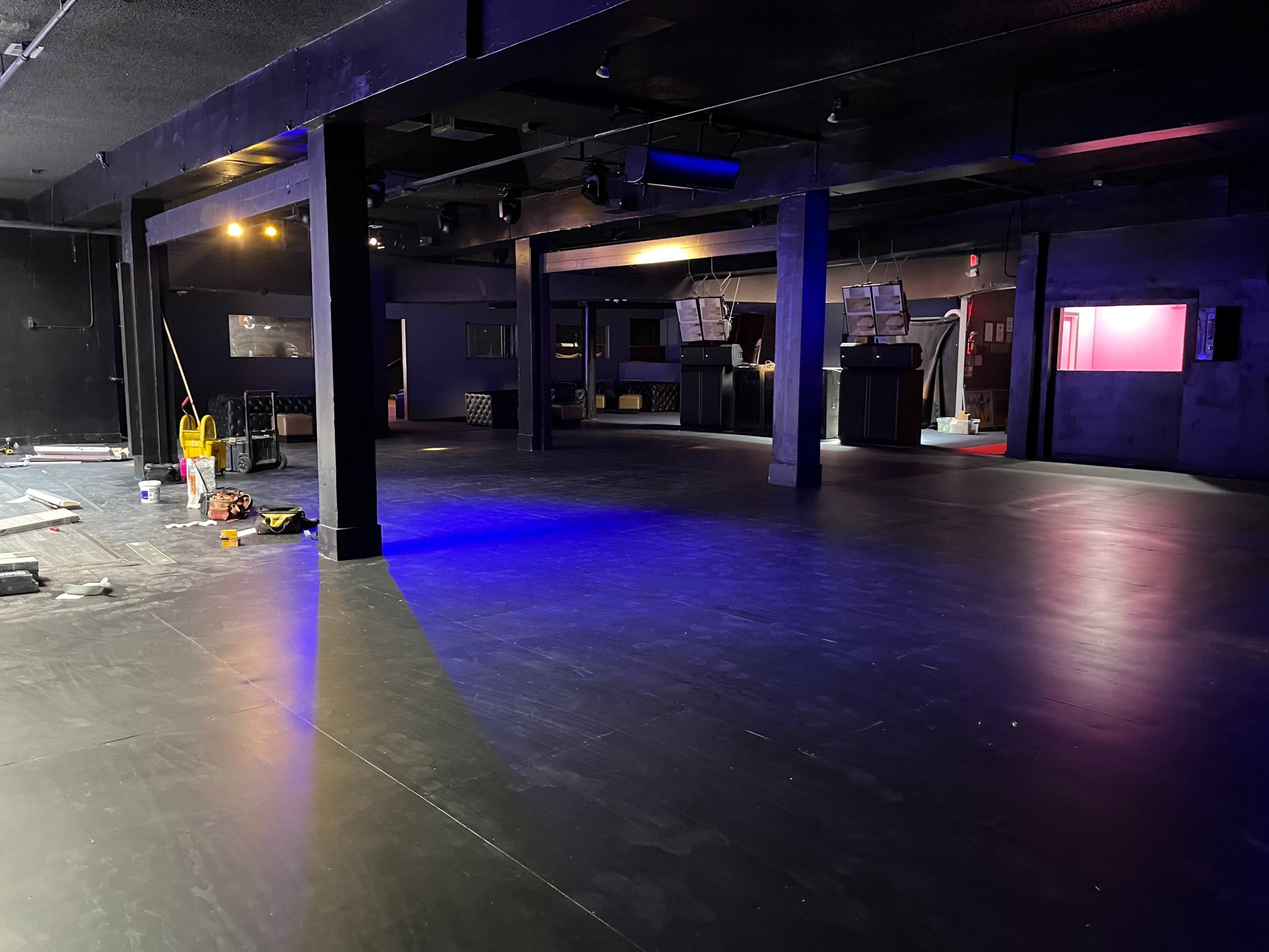 A spacious, dimly lit interior of a nightclub with black flooring and some construction tools scattered on the ground.