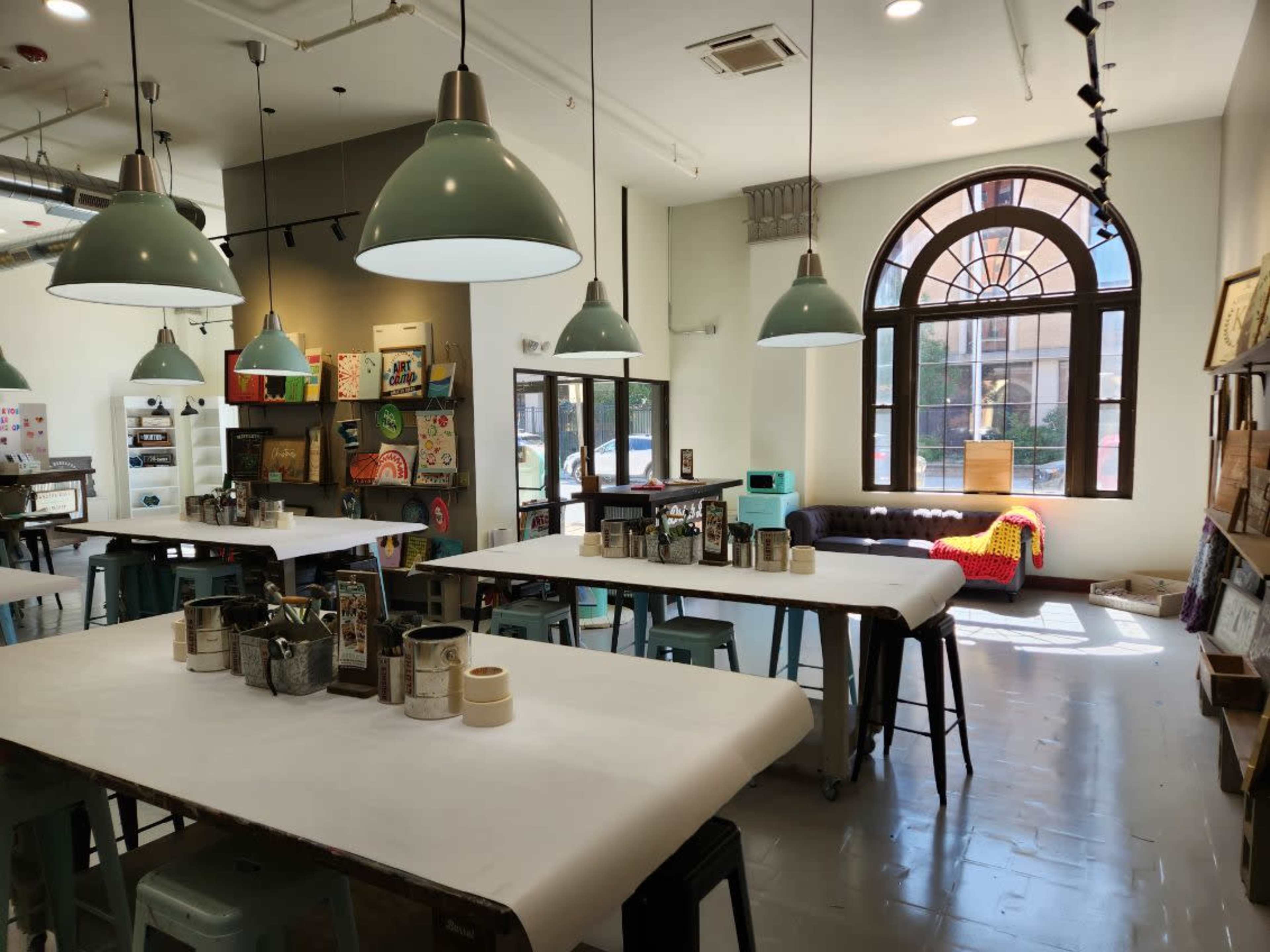 A bright art studio with large tables covered in white paper, industrial-style green pendant lights, and a cozy seating area featuring a couch near a large window.