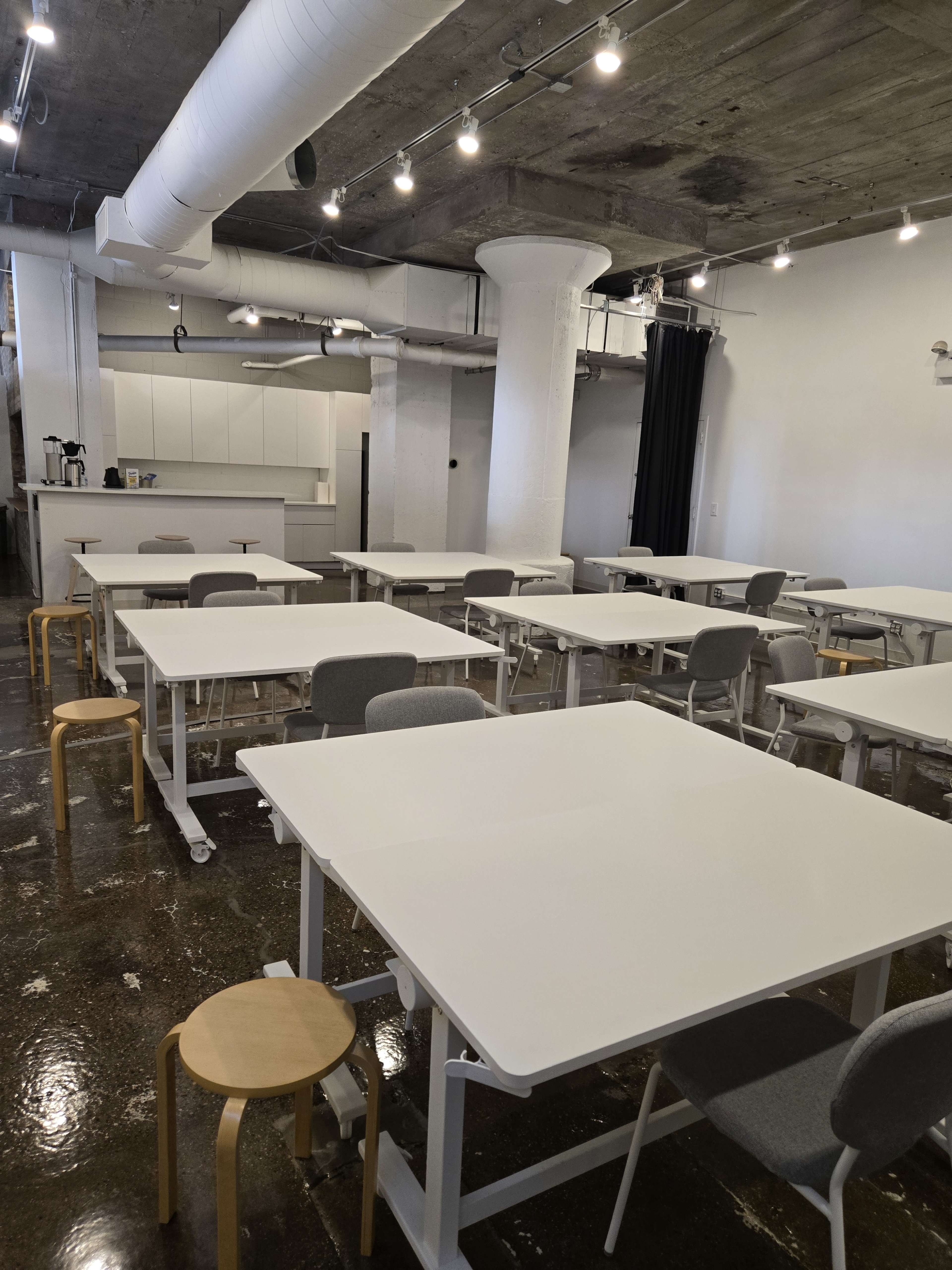 The image shows a spacious, modern classroom or workshop area with multiple white tables and gray chairs arranged in rows, along with a kitchenette in the background.