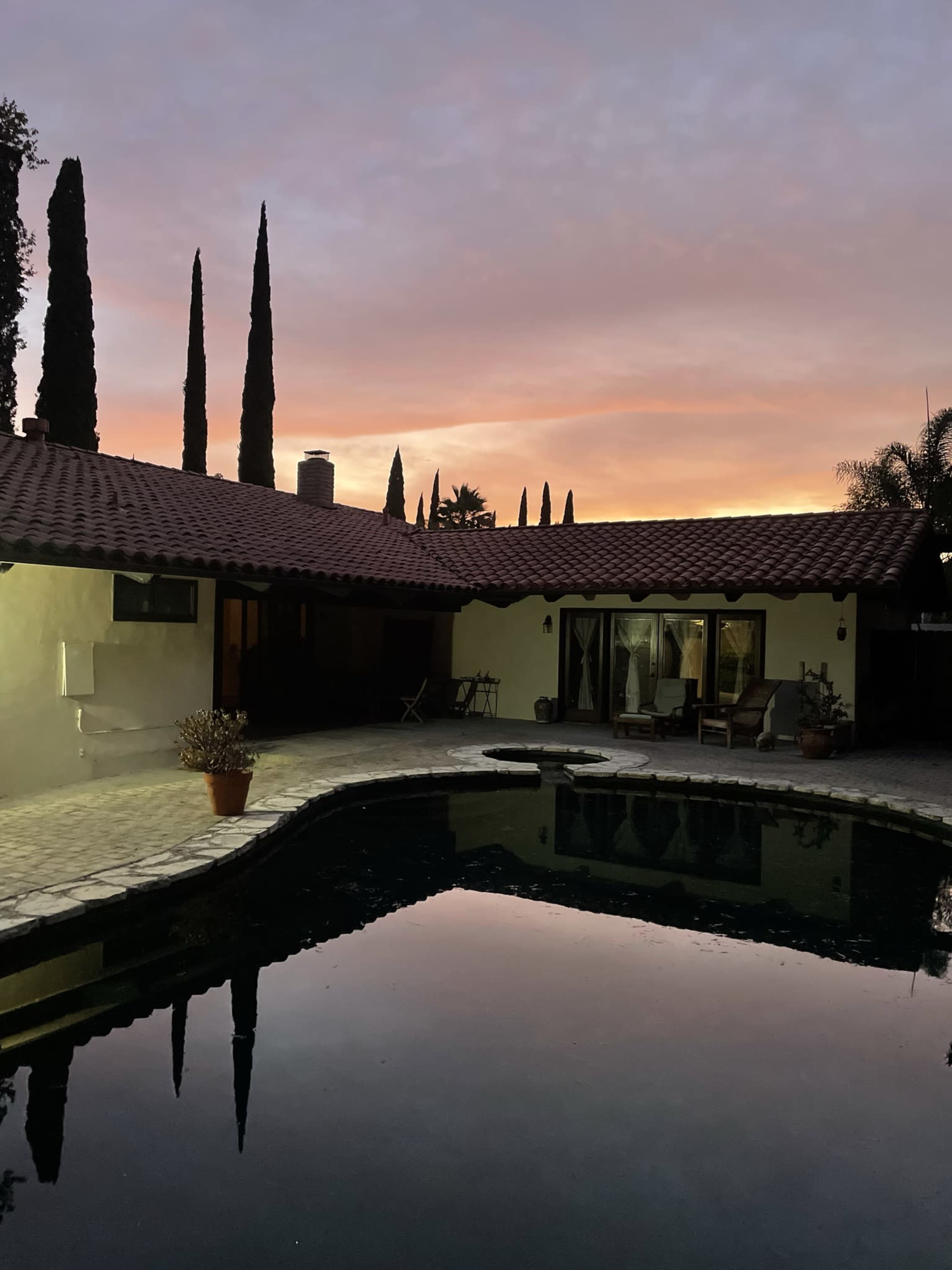 A serene backyard features a swimming pool reflecting the colorful sky at dusk, surrounded by tall cypress trees and a Spanish-style house.