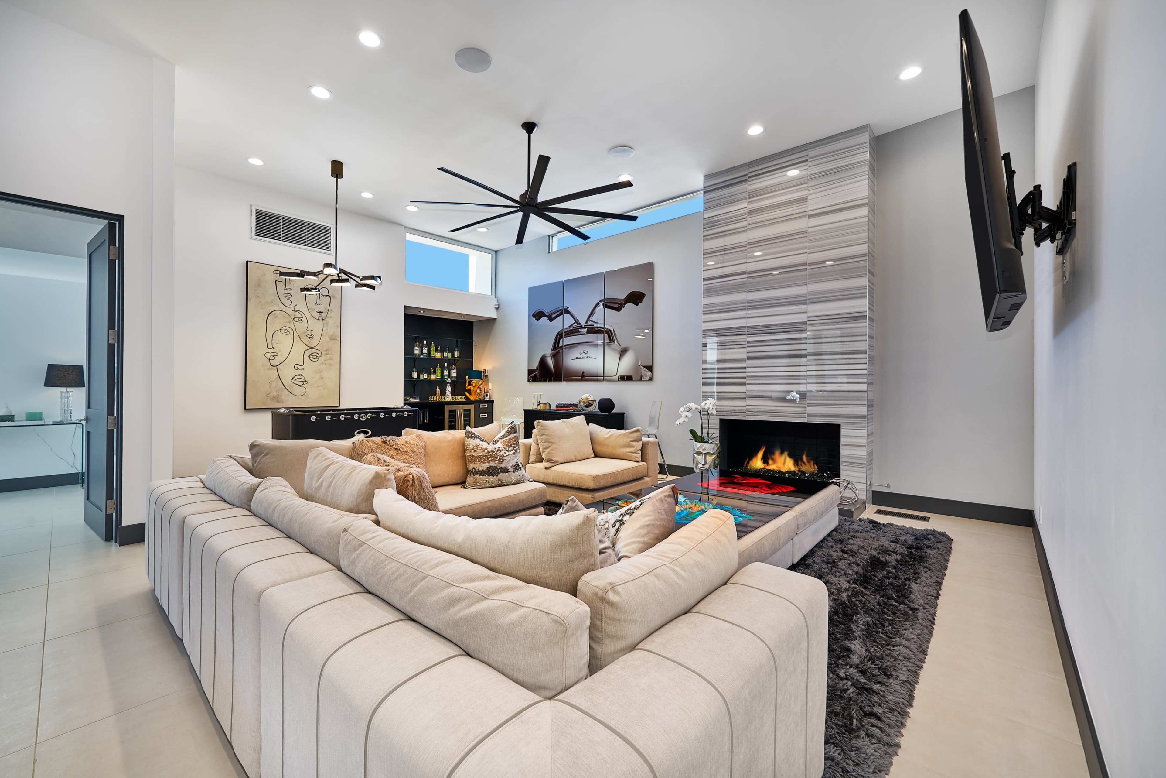 A modern living room with a sectional sofa, a wall-mounted television, a decorative fireplace, and large windows allowing natural light to enter.
