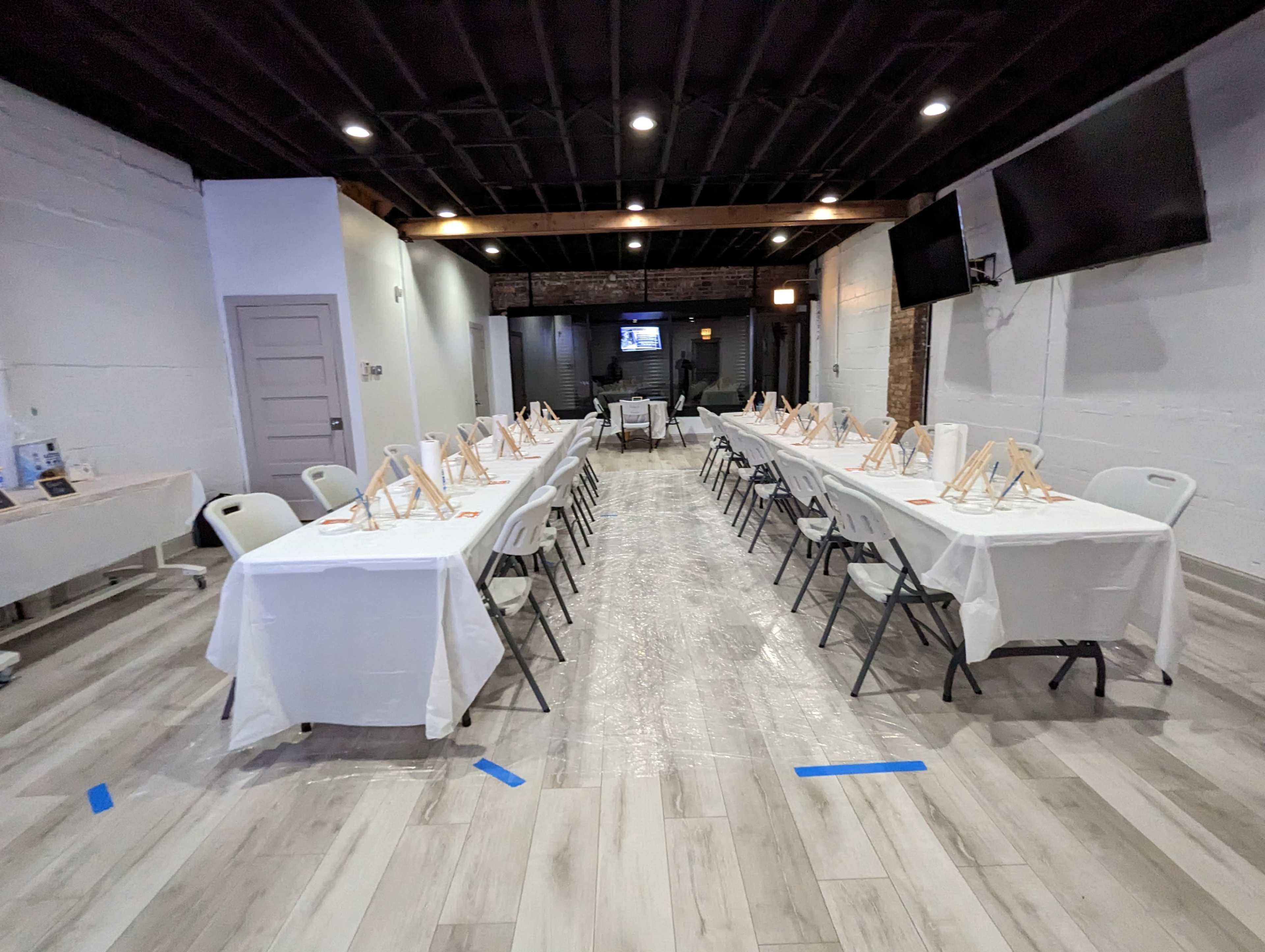 A spacious room set up for an event, with two long tables covered in white cloths and chairs arranged on either side, each table featuring an easel.