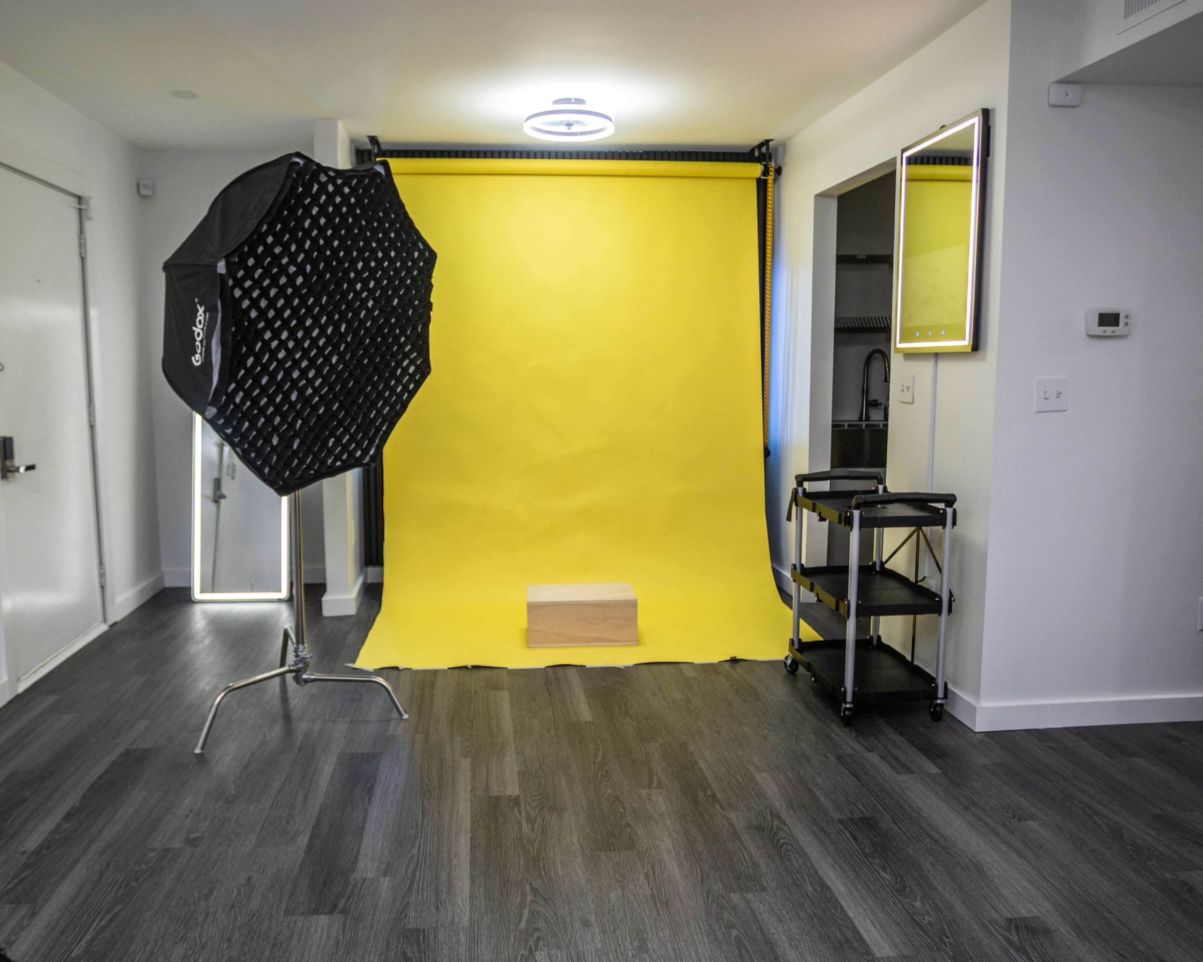 The image shows a photo studio setup with a yellow backdrop, a light umbrella, a wooden platform, and a cart in a room with modern decor.