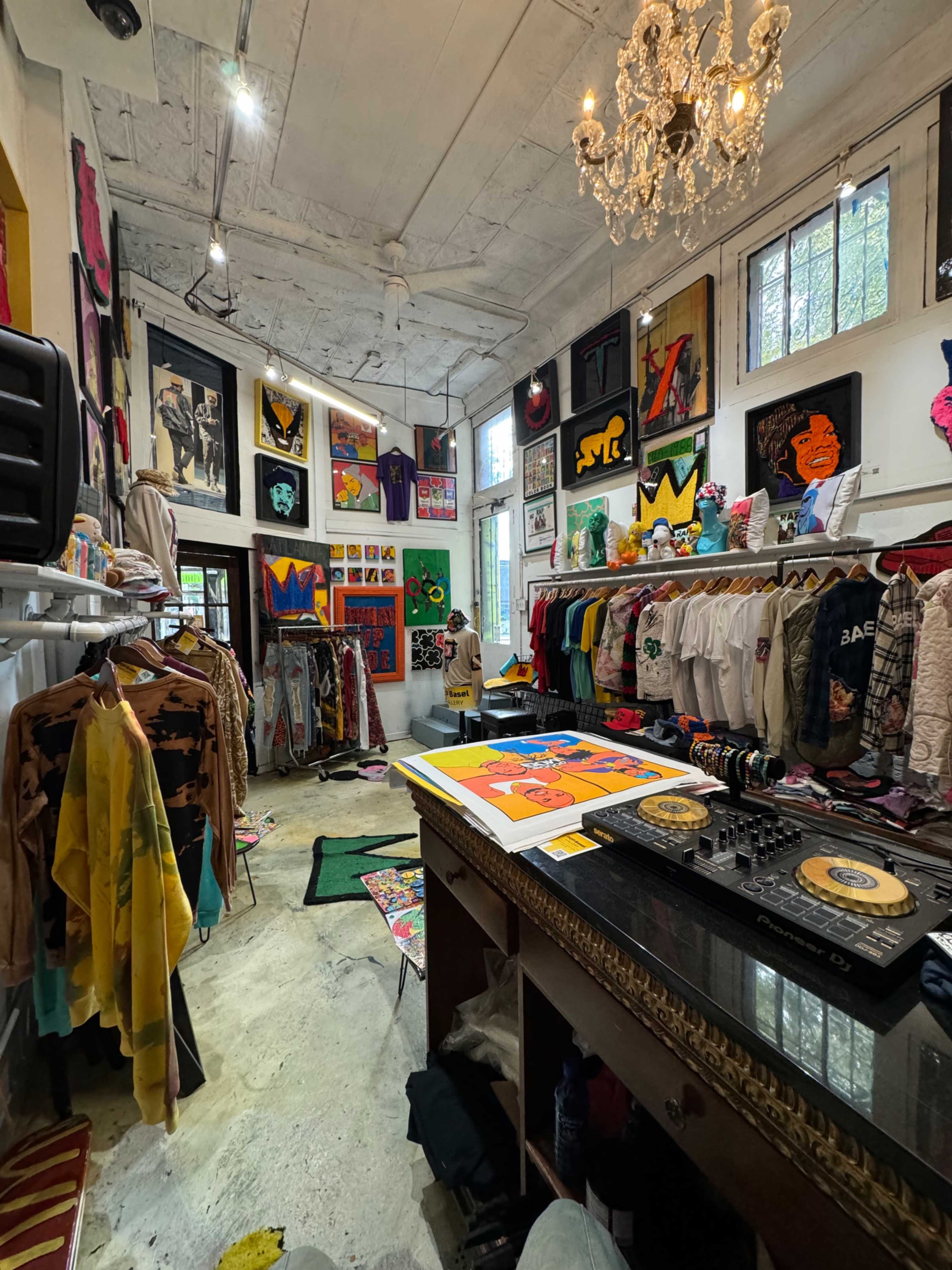 The image shows an eclectic clothing boutique with a variety of colorful apparel displayed on racks, vibrant artwork on the walls, and a DJ setup on a counter.