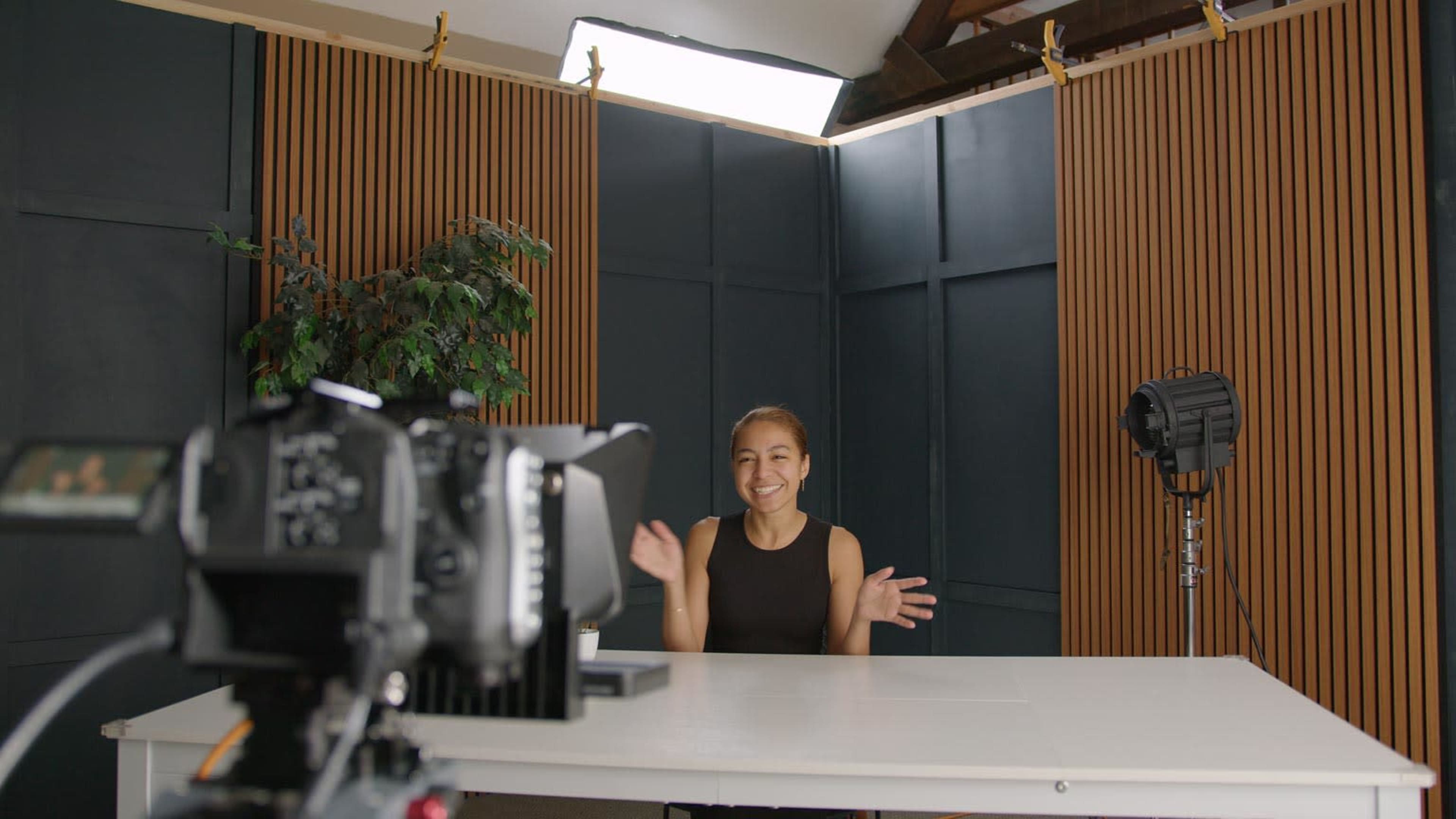 A woman is sitting at a table in a studio setting, smiling and waving at a camera positioned in front of her.