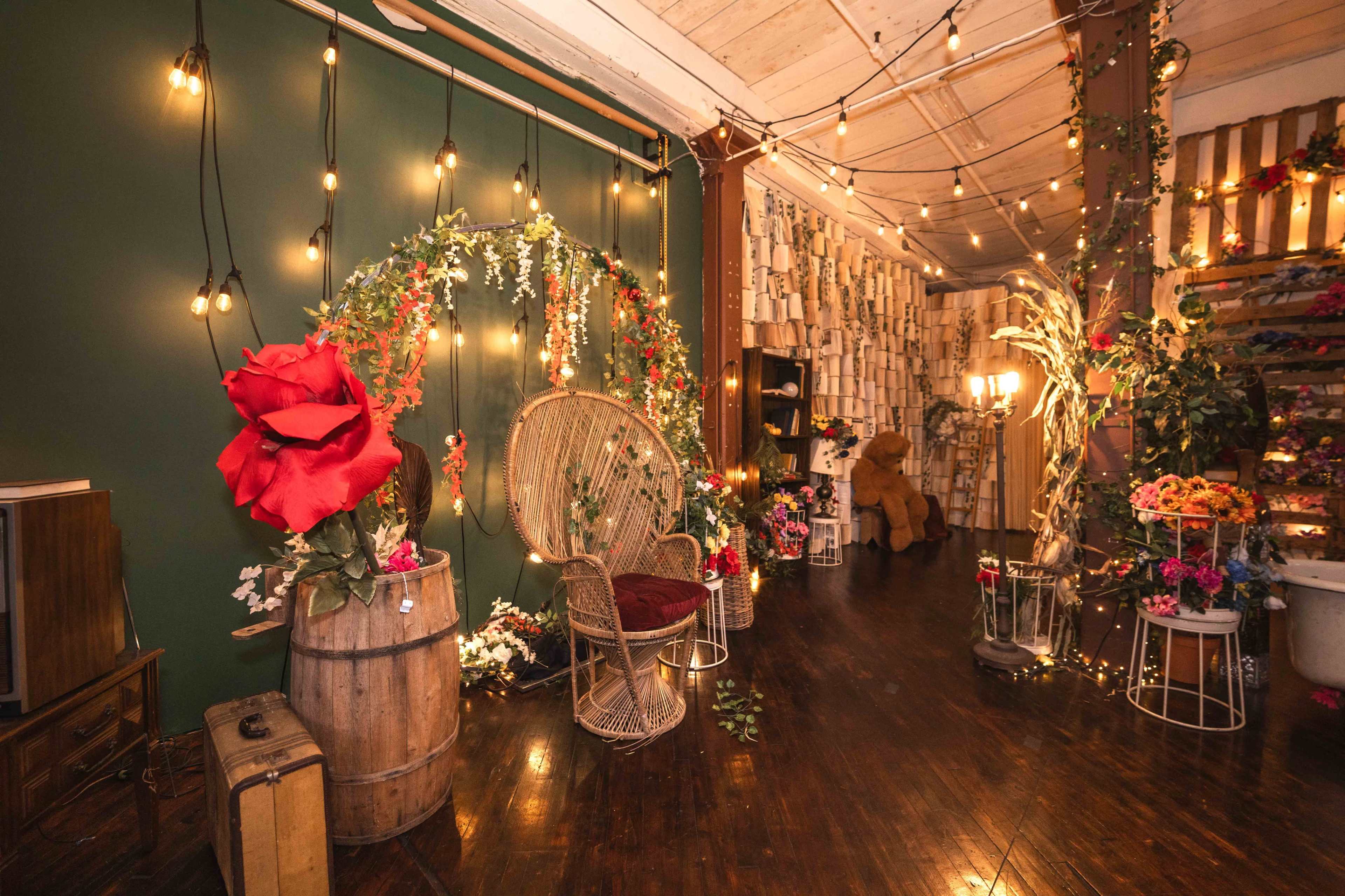 The image shows an indoor space decorated with fairy lights, floral arrangements, and a wicker chair surrounded by various props and a wooden backdrop.
