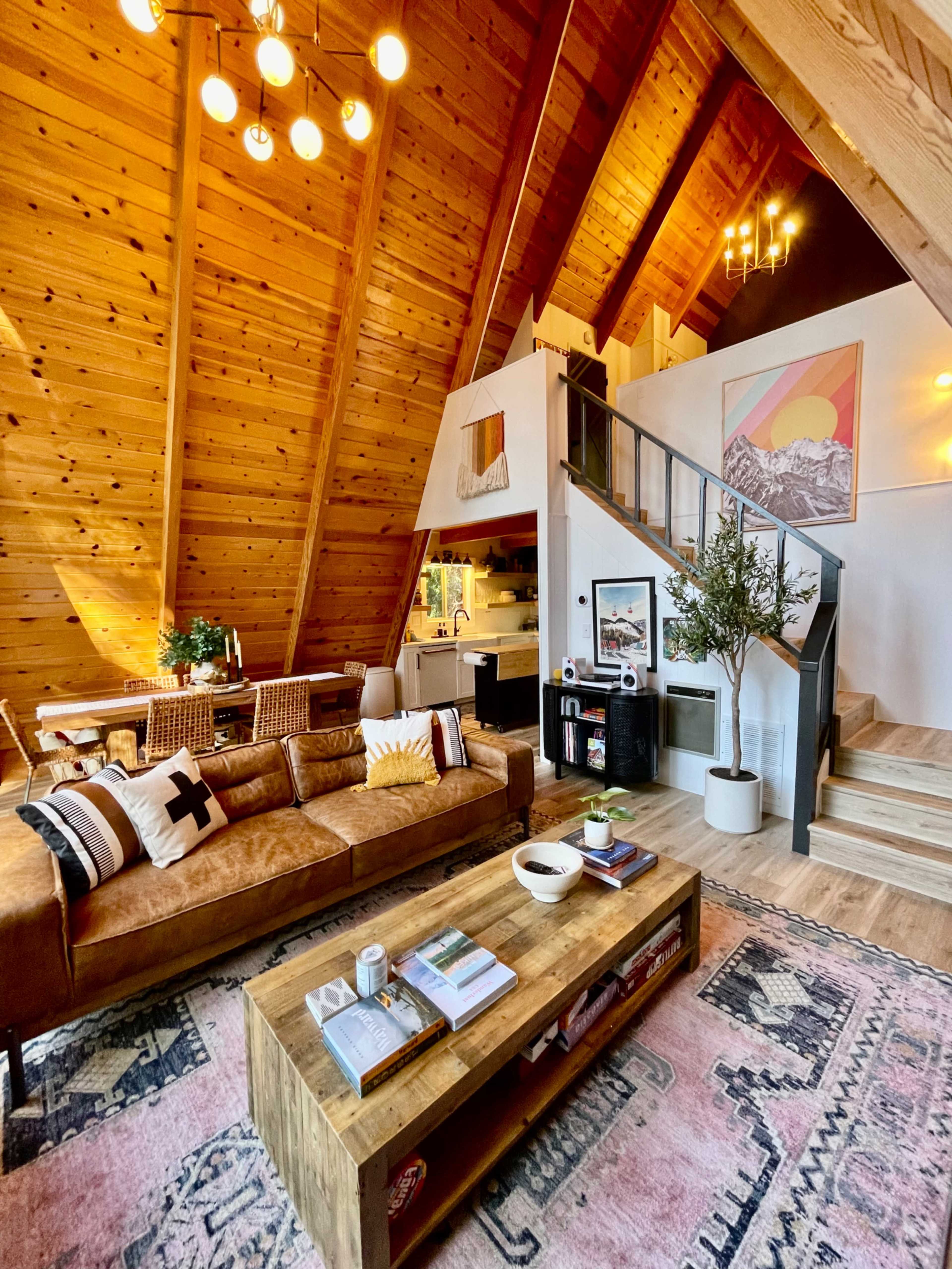A cozy, wooden A-frame living room featuring a brown leather sofa, a large coffee table, and a staircase leading to the upper level.
