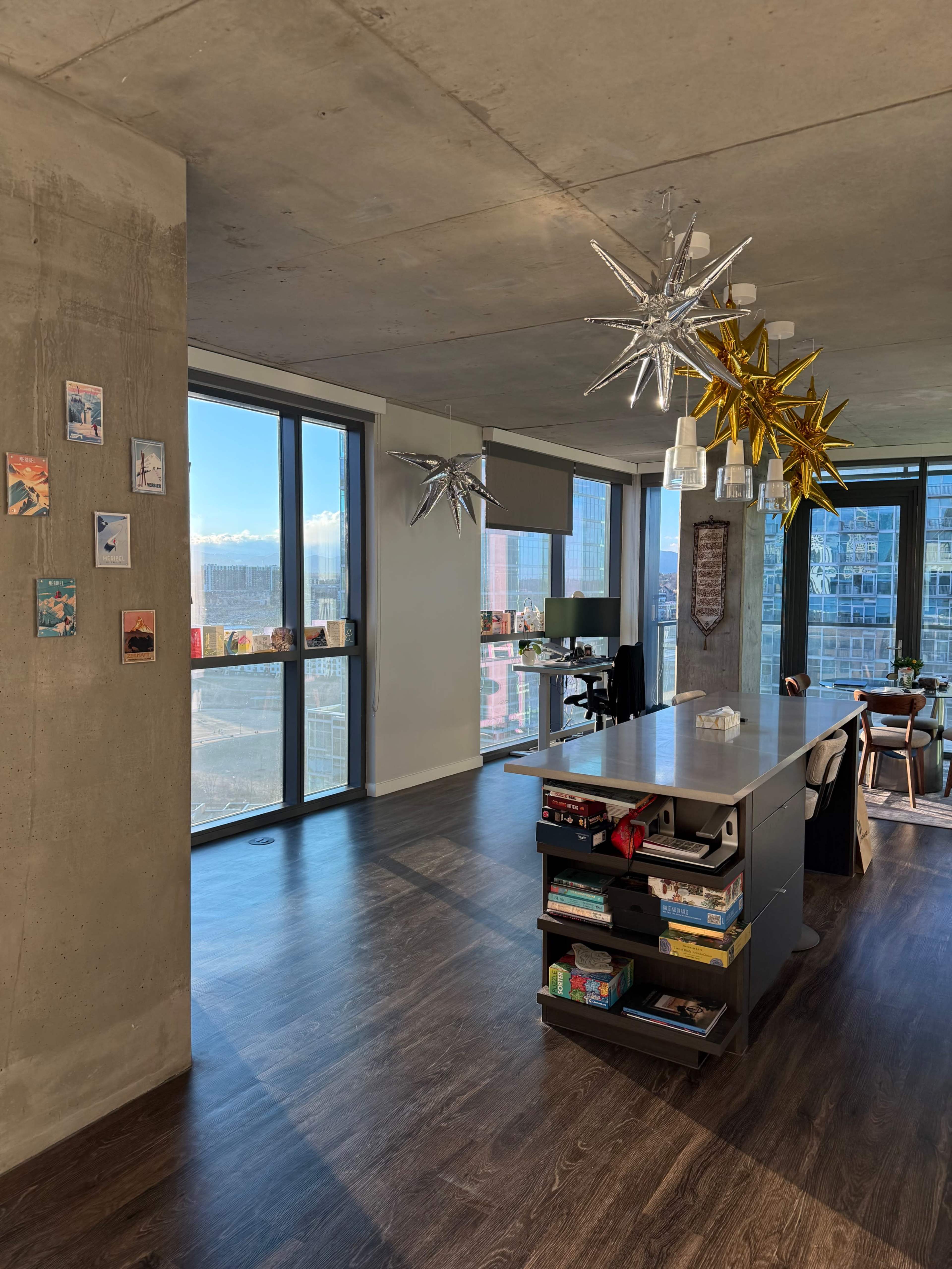 The image shows a modern, open-concept apartment with large windows displaying a cityscape and decorative star-shaped lights hanging from the ceiling.
